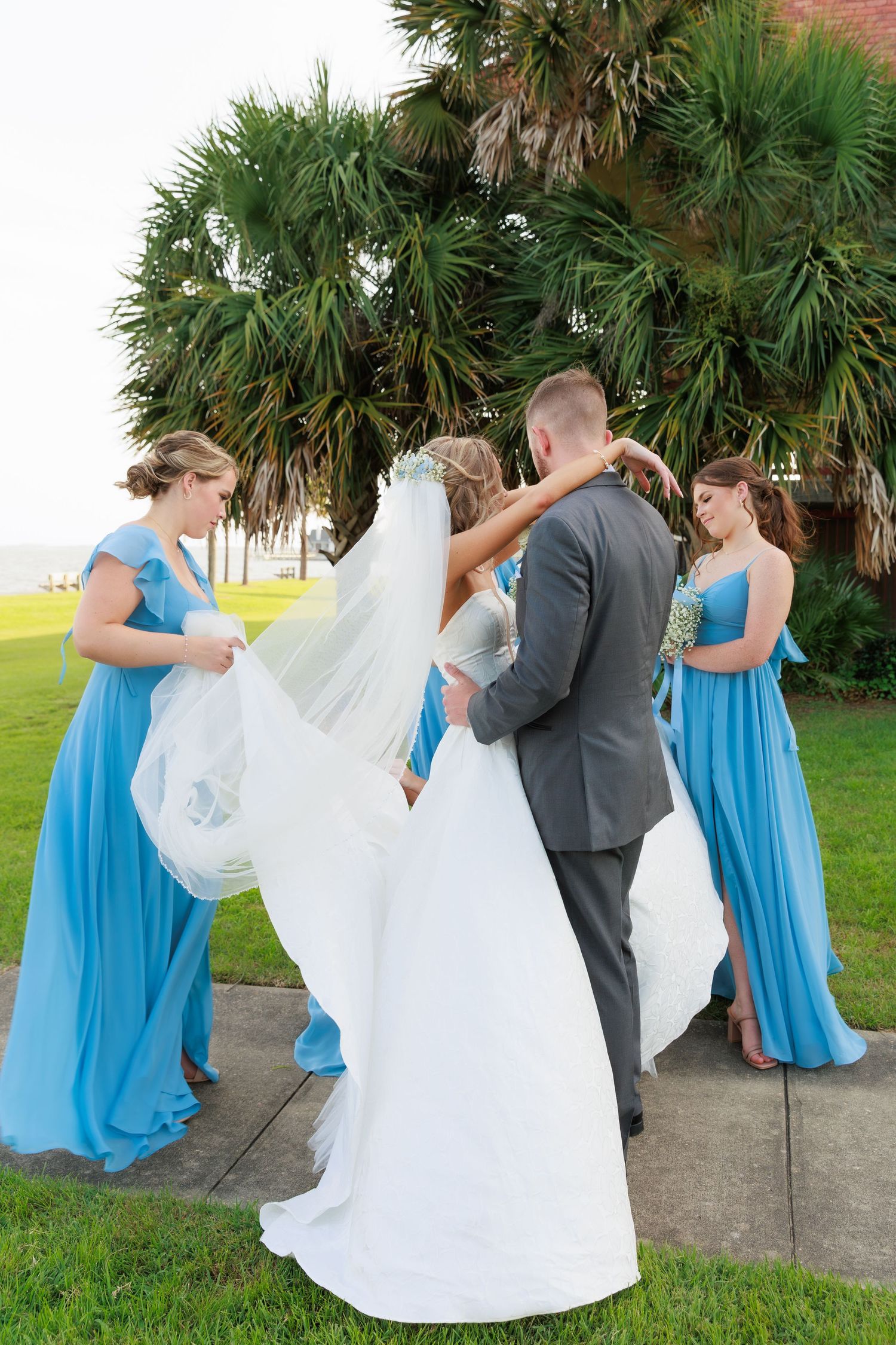 Bridesmaids in blue dresses assist with bride's veil during outdoor wedding photo.