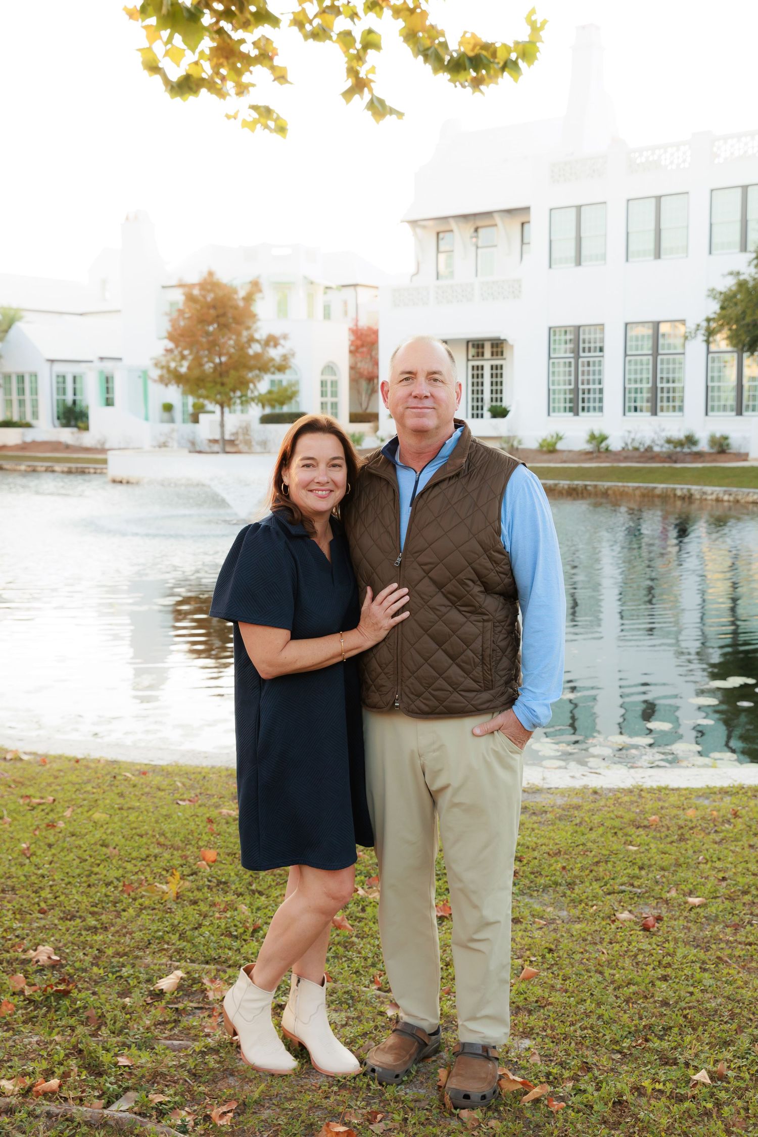 Couple poses together on grass near a white building with water feature in background.