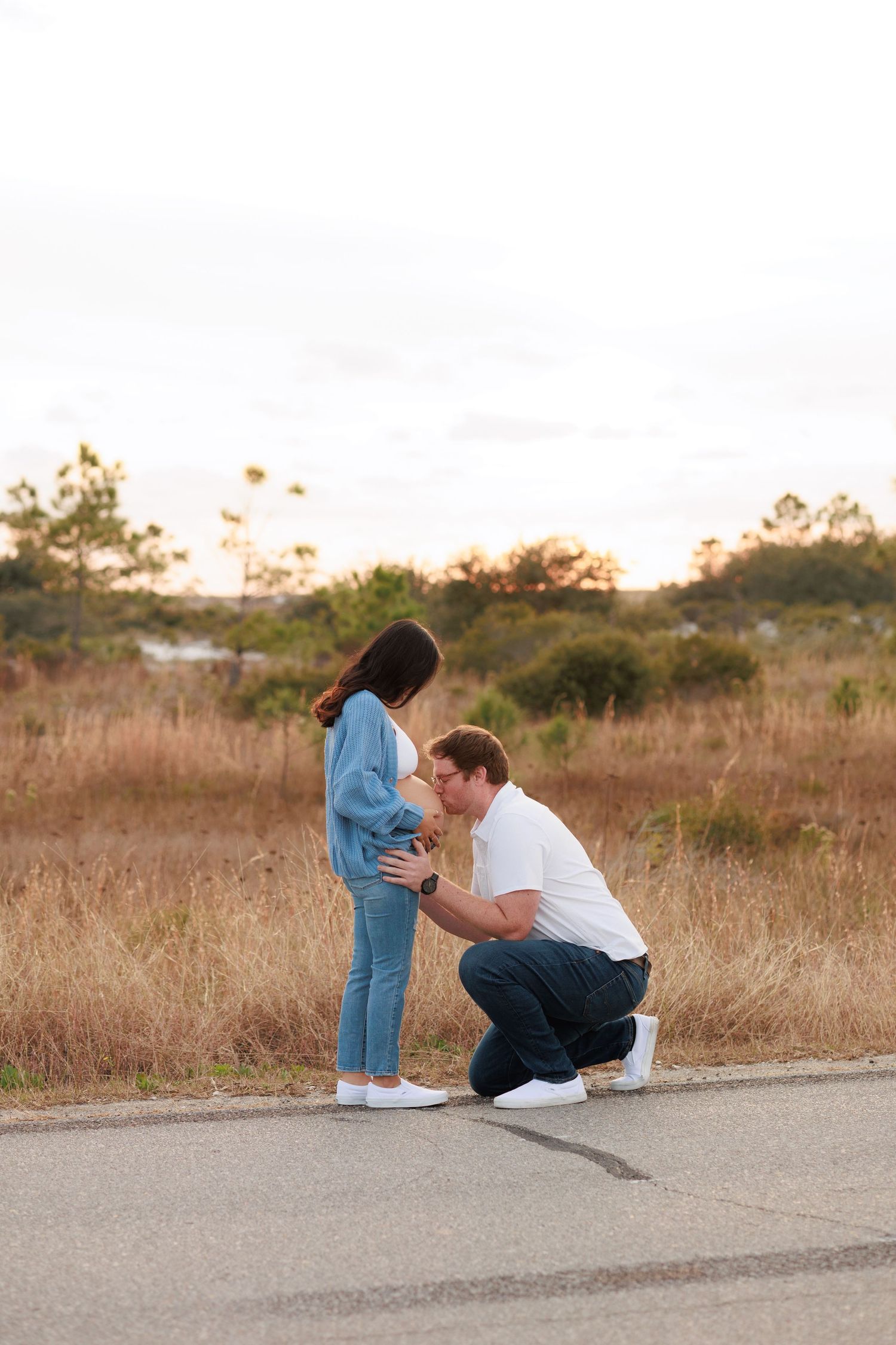A couple shares a tender moment while sitting together on a concrete path at sunset in a grassy field.