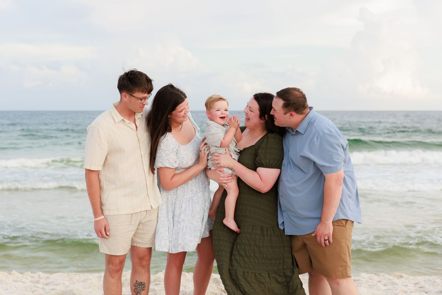A group poses together for casual family photos on a sunny beach with ocean waves in the background.