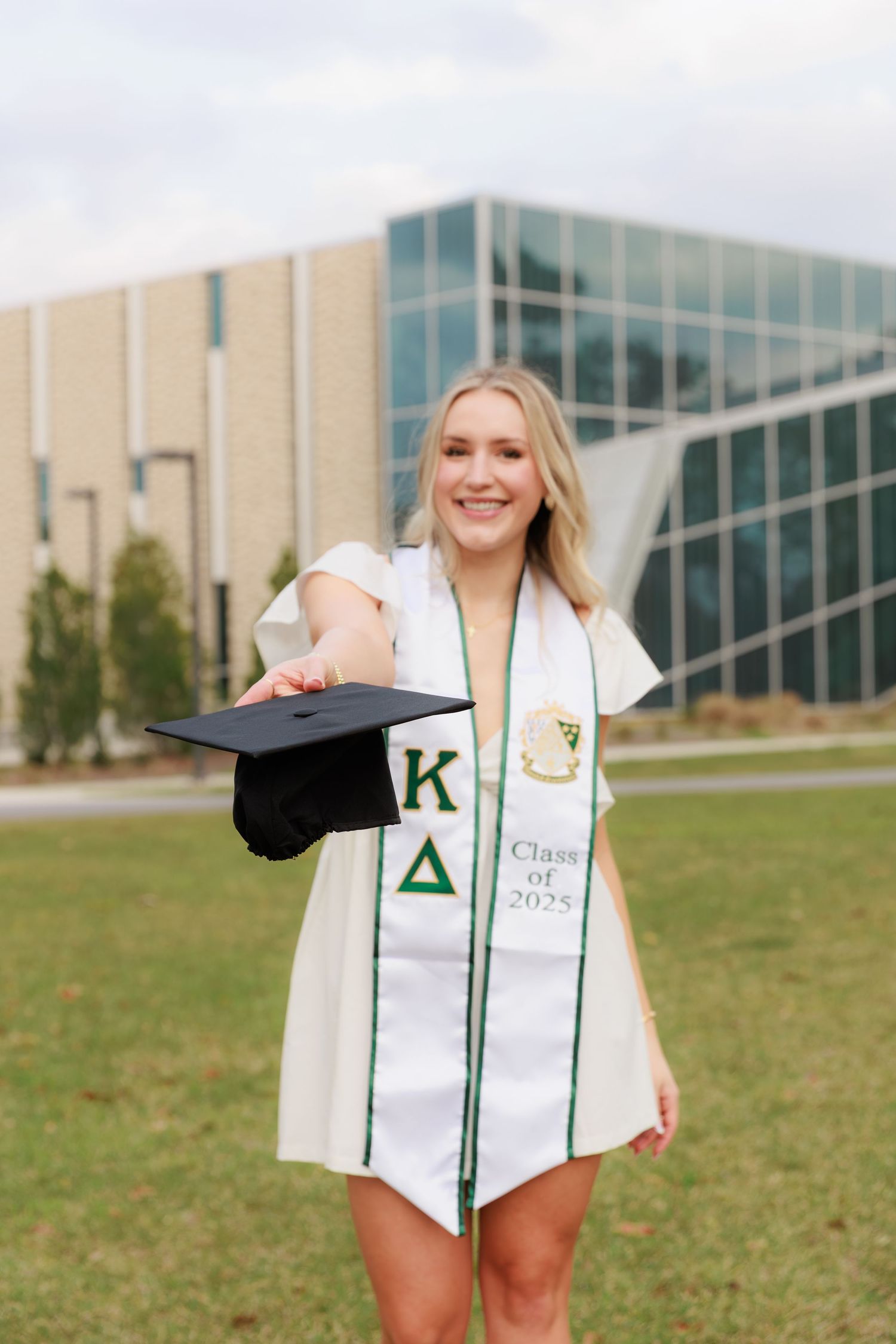 Smiling blonde graduate holds cap toward camera wearing Kappa Delta sash reading Class of 2025 outdoors on campus.