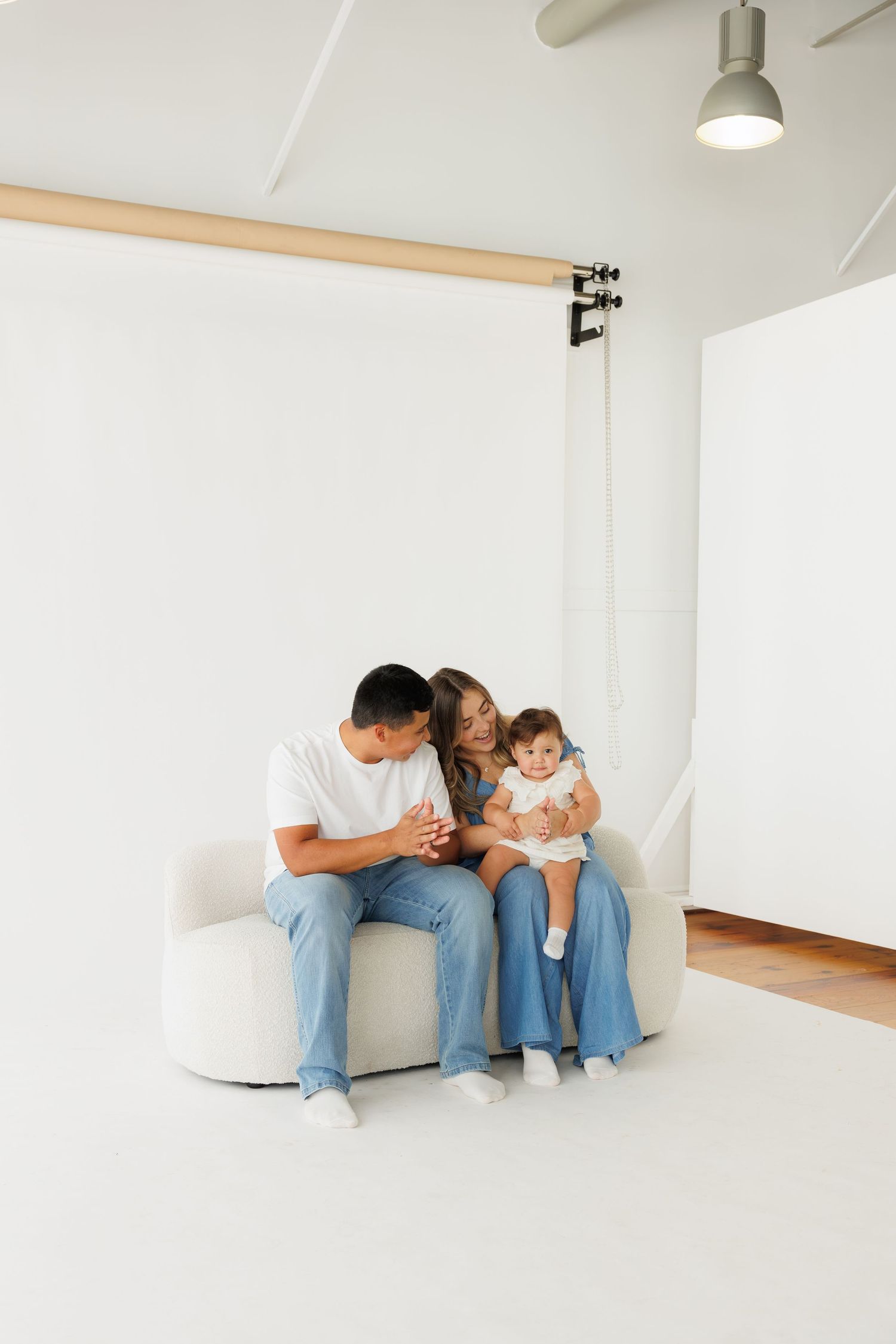 Young family with baby sitting on white sofa in bright studio, parents smiling at infant in denim outfits.
