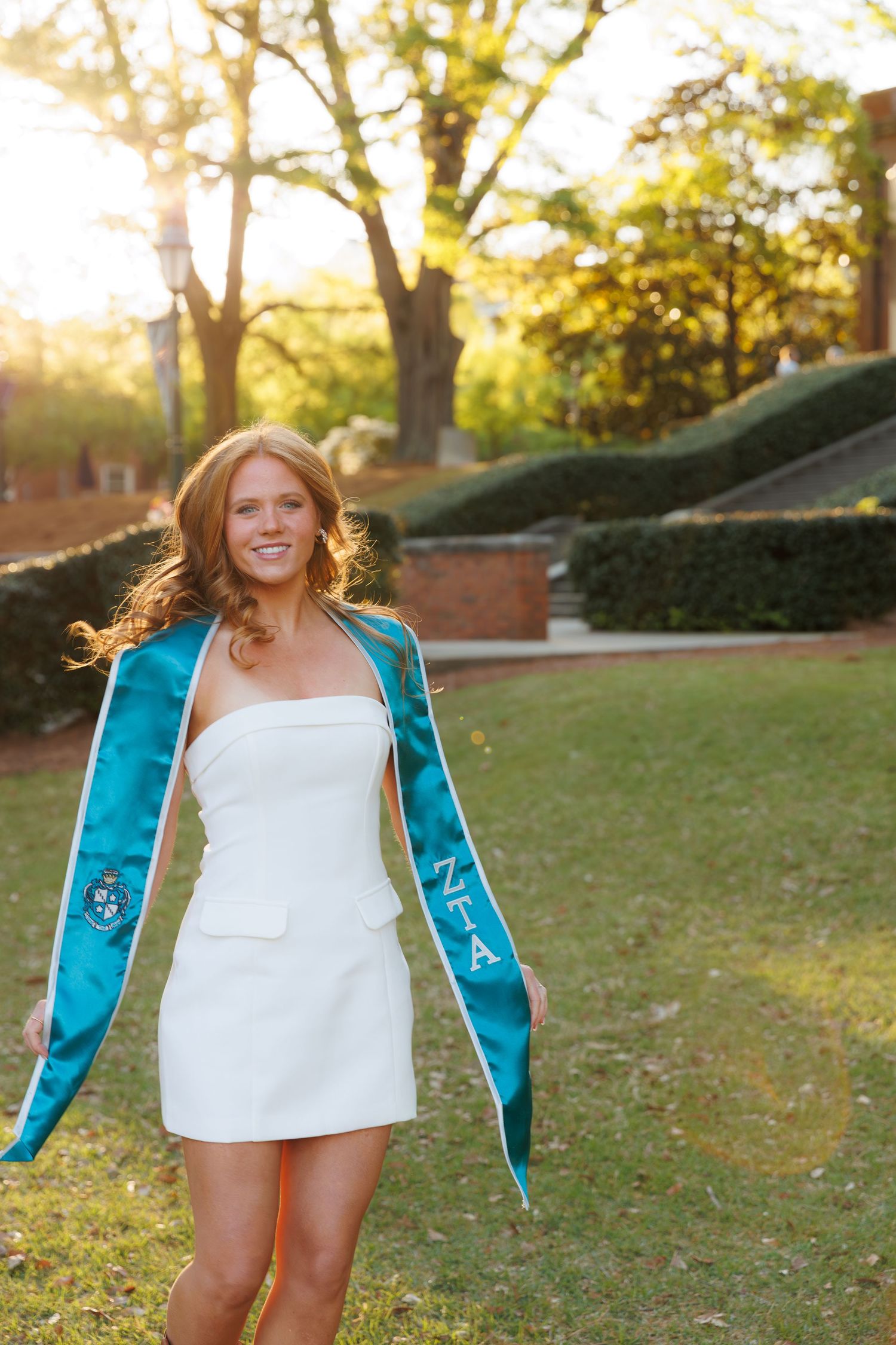 Smiling young woman in white mini dress wearing teal graduation stole outdoors on sunny campus grounds.