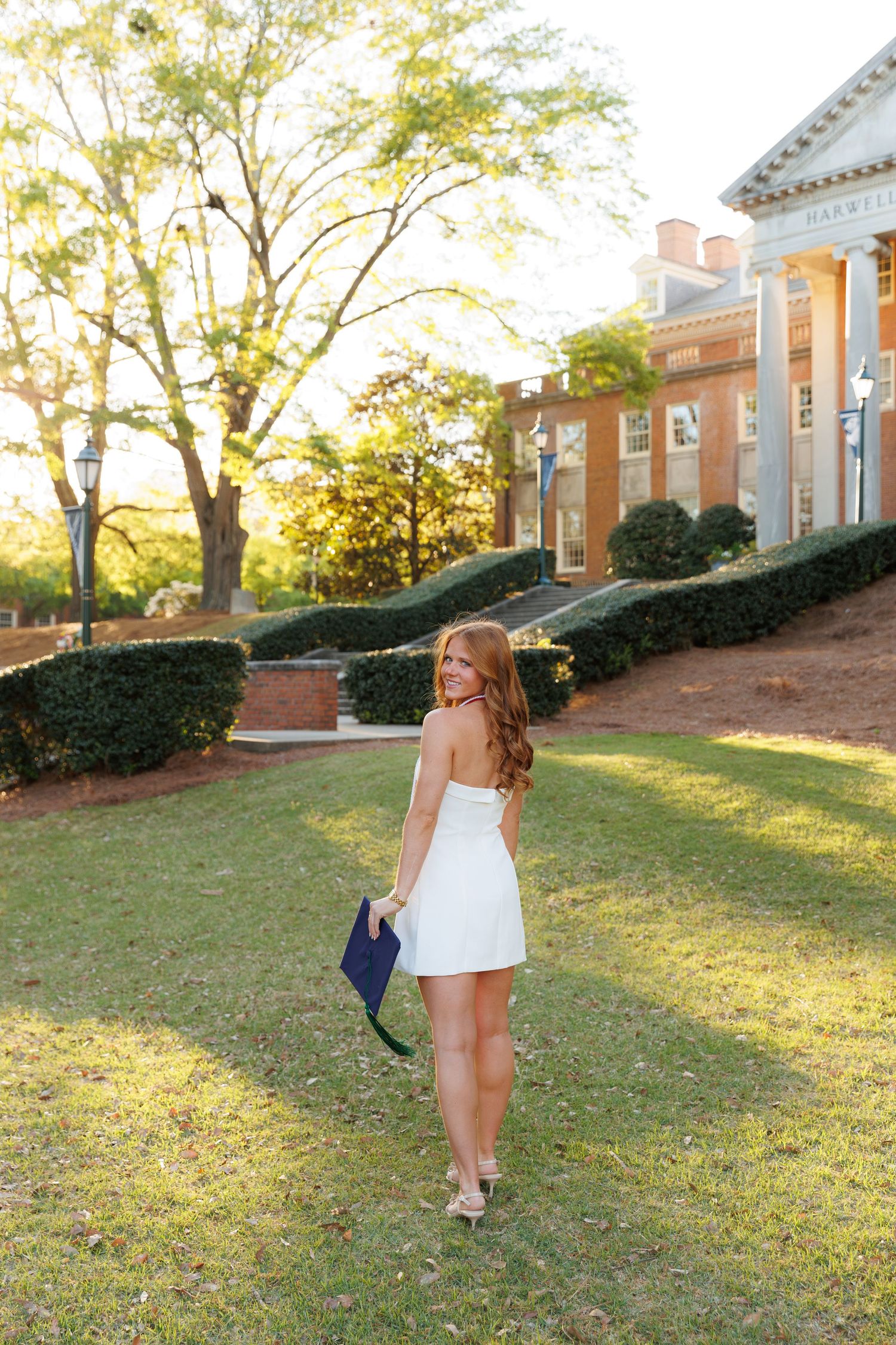 Young woman in white dress holding graduation cap walks on green campus lawn near brick building at golden hour.