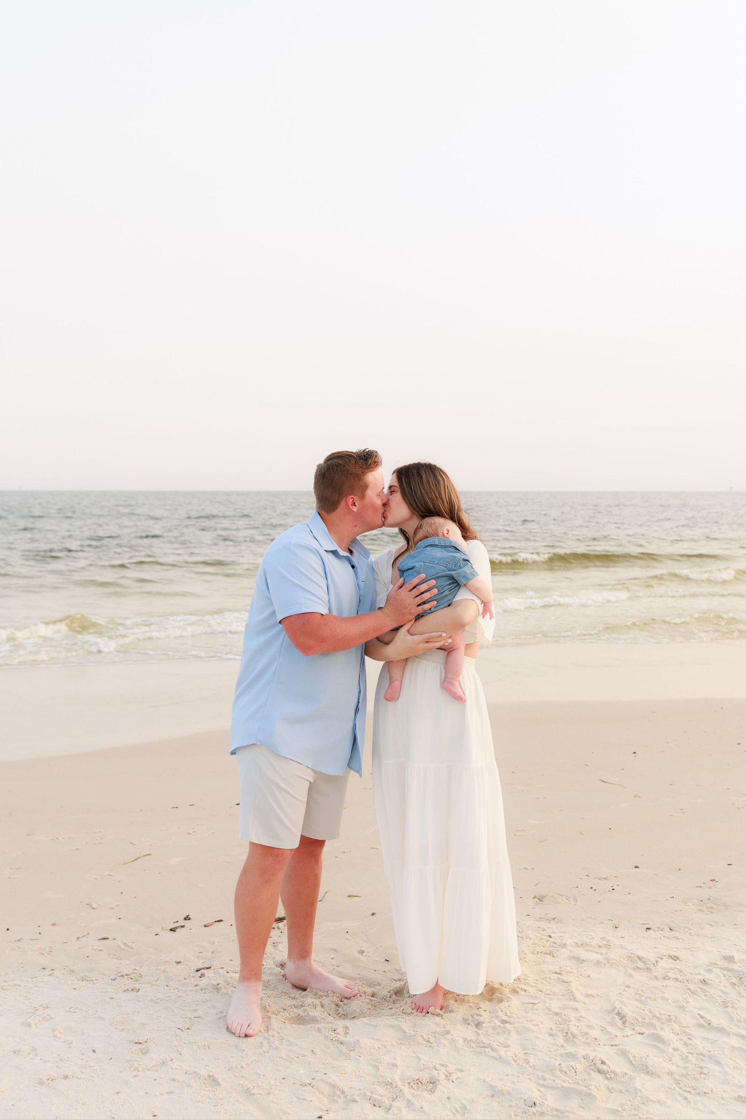 A couple sharing a kiss on a sandy beach while holding their baby, with ocean waves in the background.