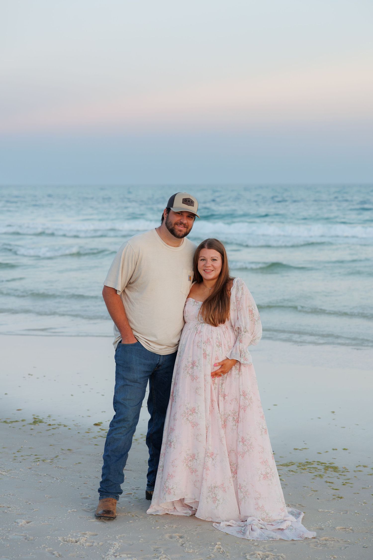 Smiling couple standing on beach at sunset, woman in floral maternity dress, man in jeans and trucker hat.
