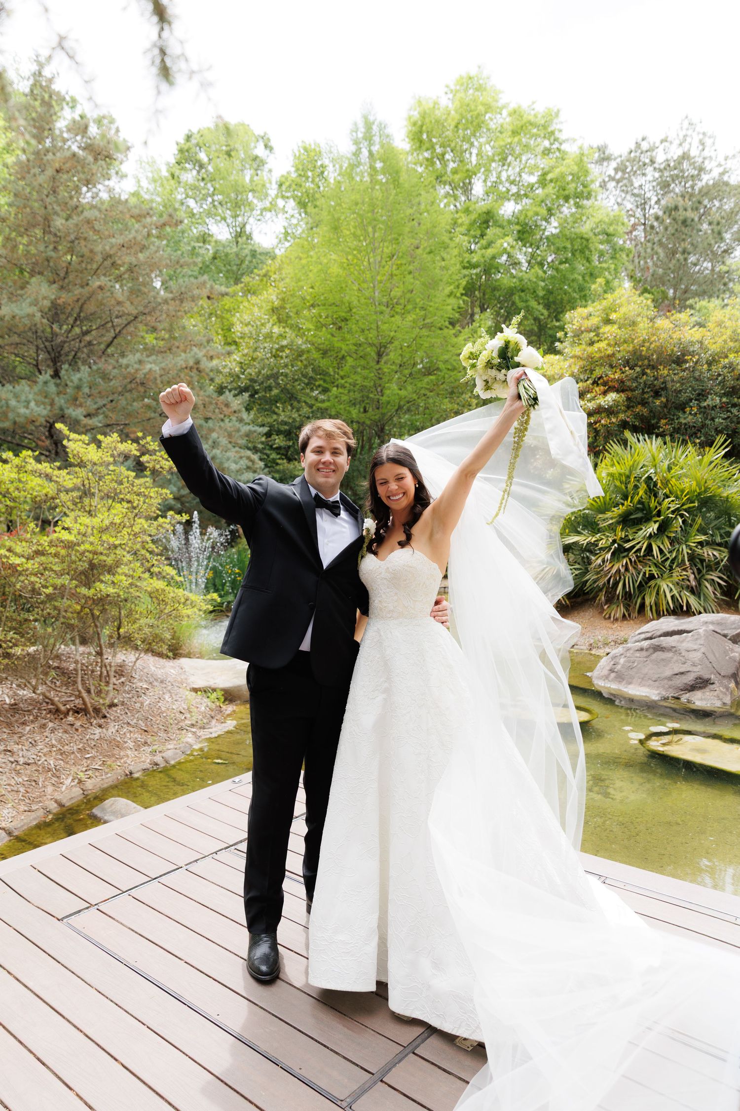 Joyful newlyweds celebrate on a wooden deck in a lush garden, groom in tuxedo and bride in white gown raising arms triumphantly.