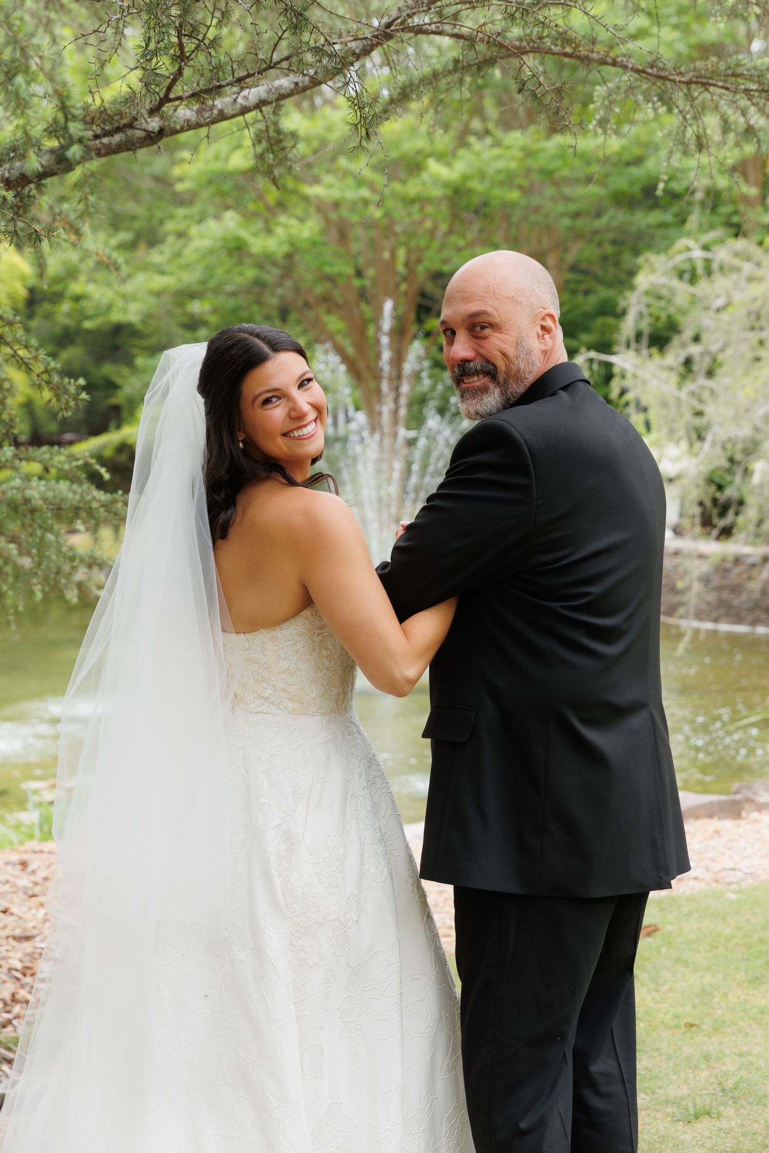 Smiling bride in white gown and veil poses with groom in black suit near a garden fountain.