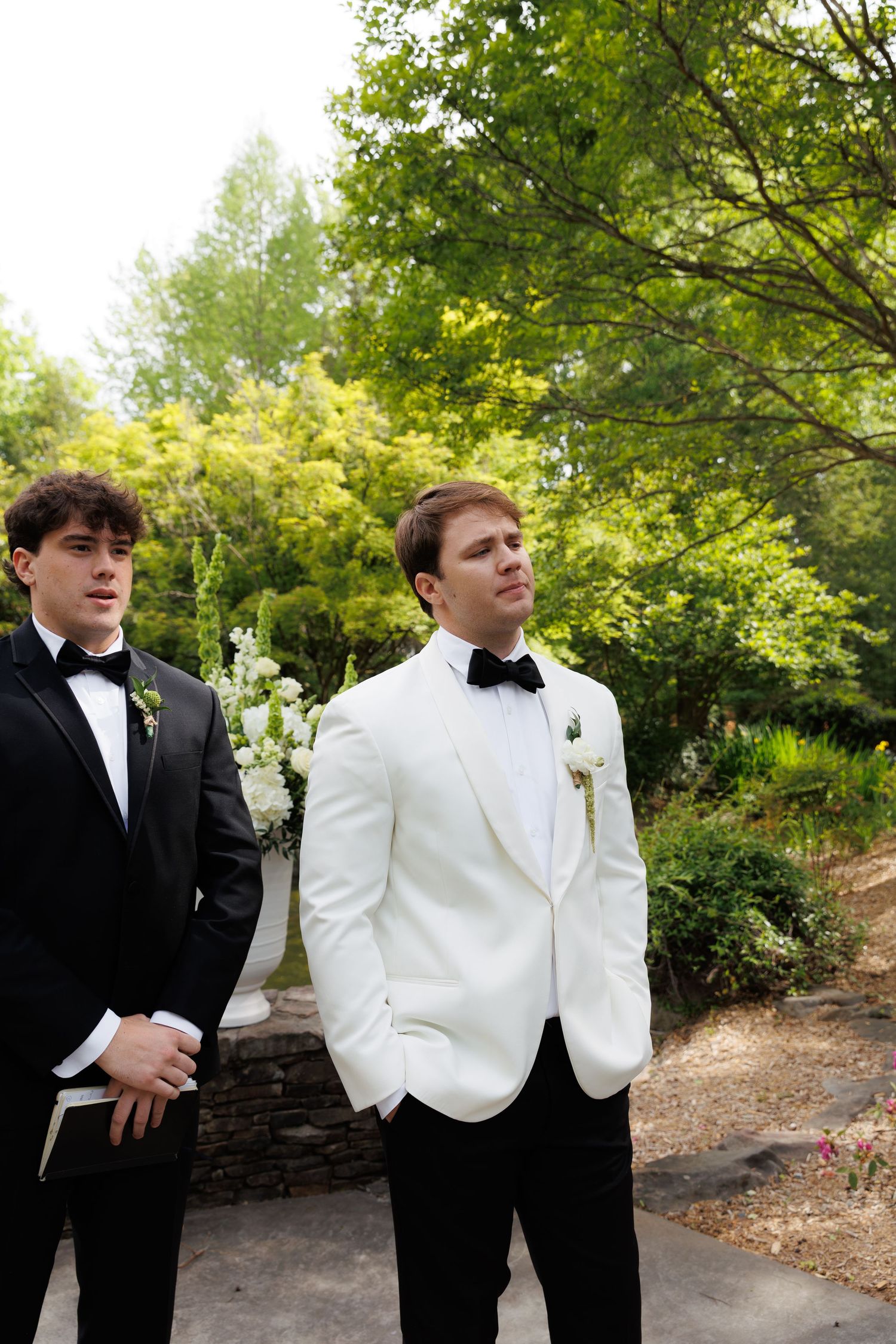 Groom in white tuxedo jacket stands with groomsman in black tux at outdoor wedding ceremony.