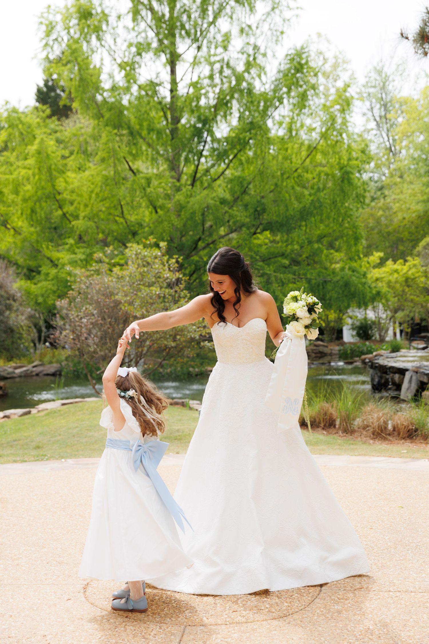 Bride in strapless white gown twirls flower girl in white dress with blue sash on outdoor patio surrounded by greenery.