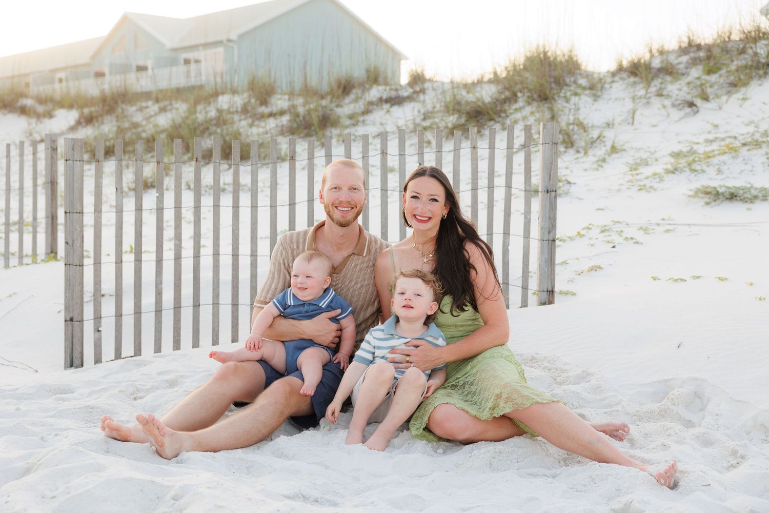 Happy family of four with two young boys sitting together on white sand beach near dunes.