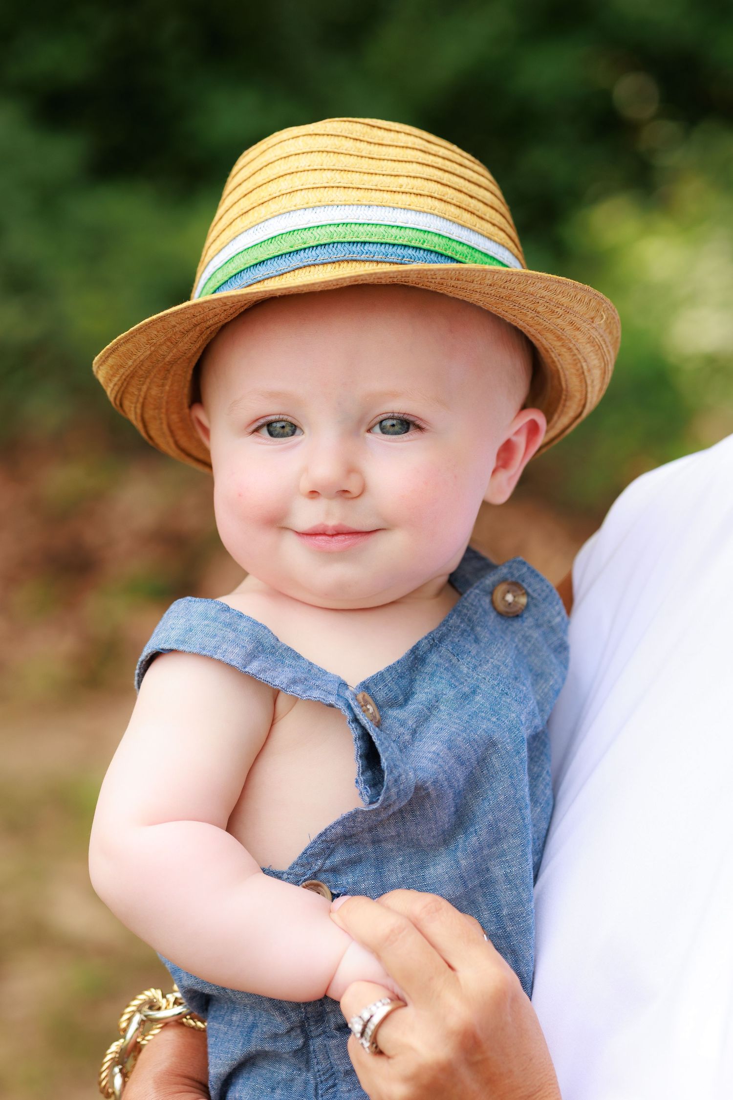 A baby wearing a straw hat with a turquoise band and denim romper smiles while being held outdoors.