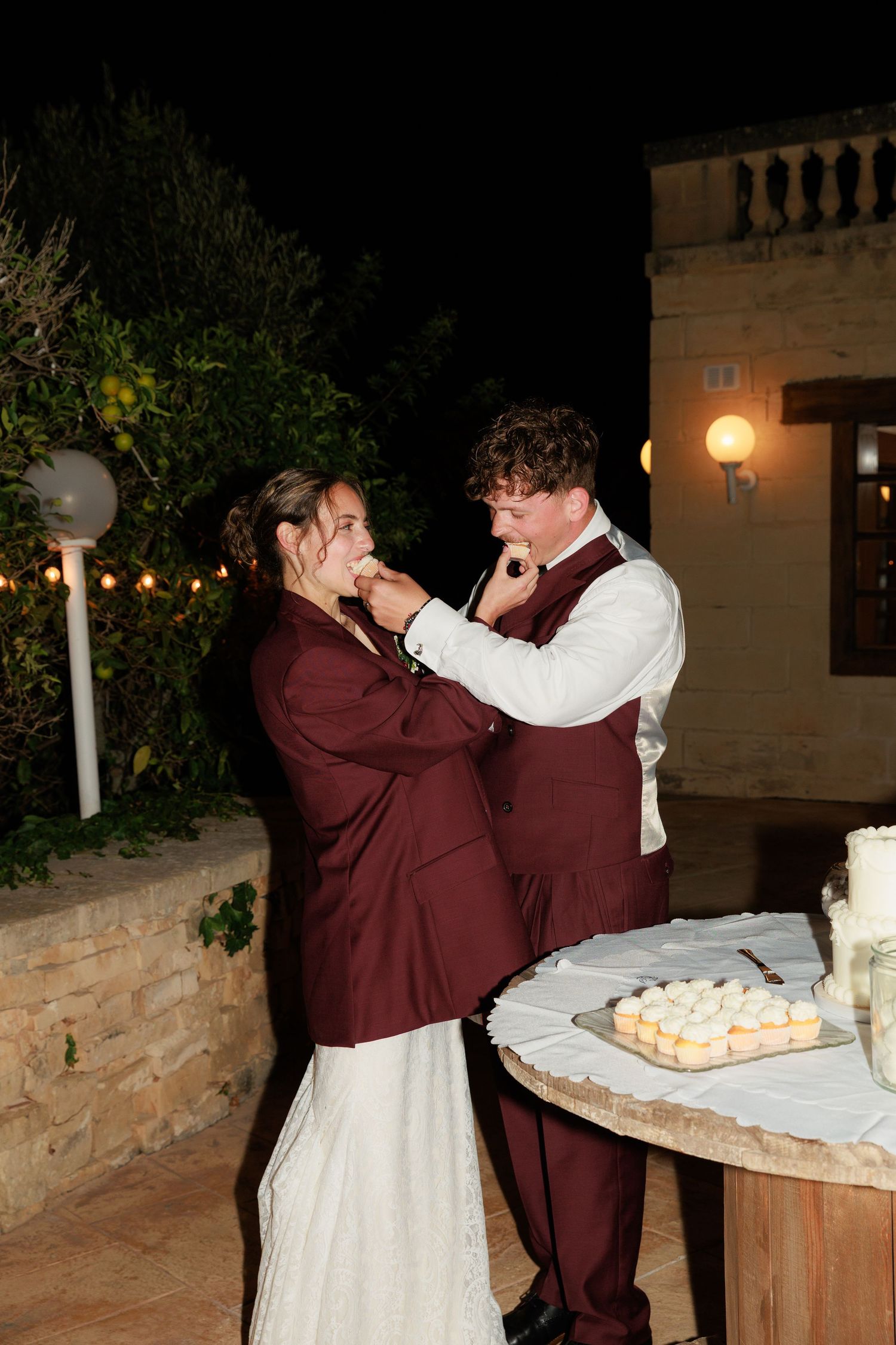 Couple in burgundy attire share a playful cake-feeding moment at their wedding reception next to a dessert table.