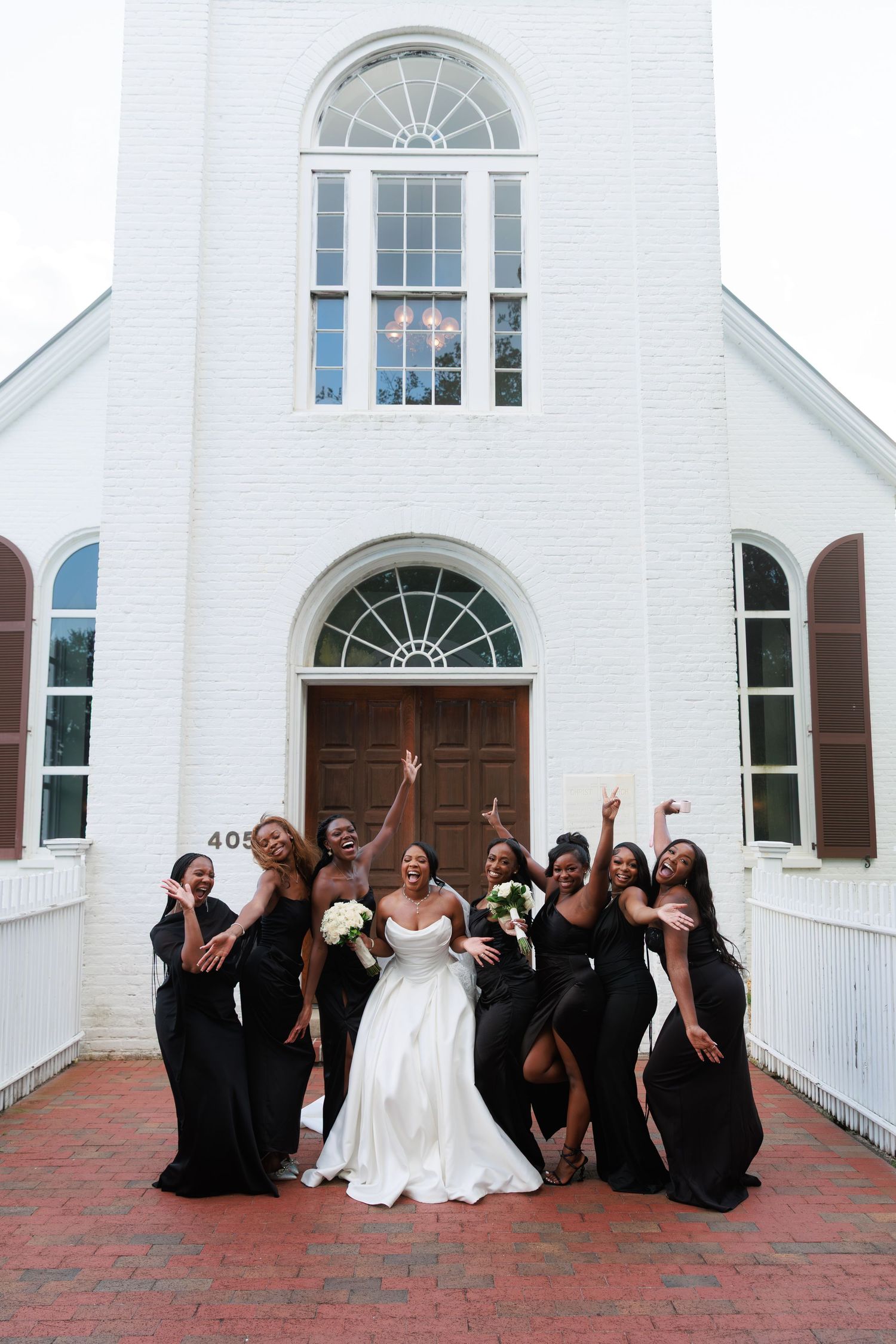 Wedding party in black dresses and white gown poses playfully in front of white church building with arched windows.