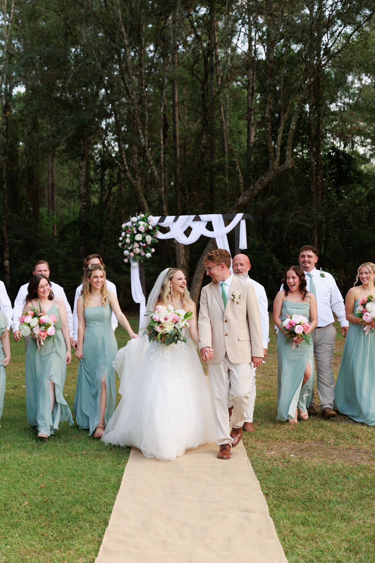 Wedding party walks together on outdoor path wearing mint green dresses and khaki suits under white draped archway.