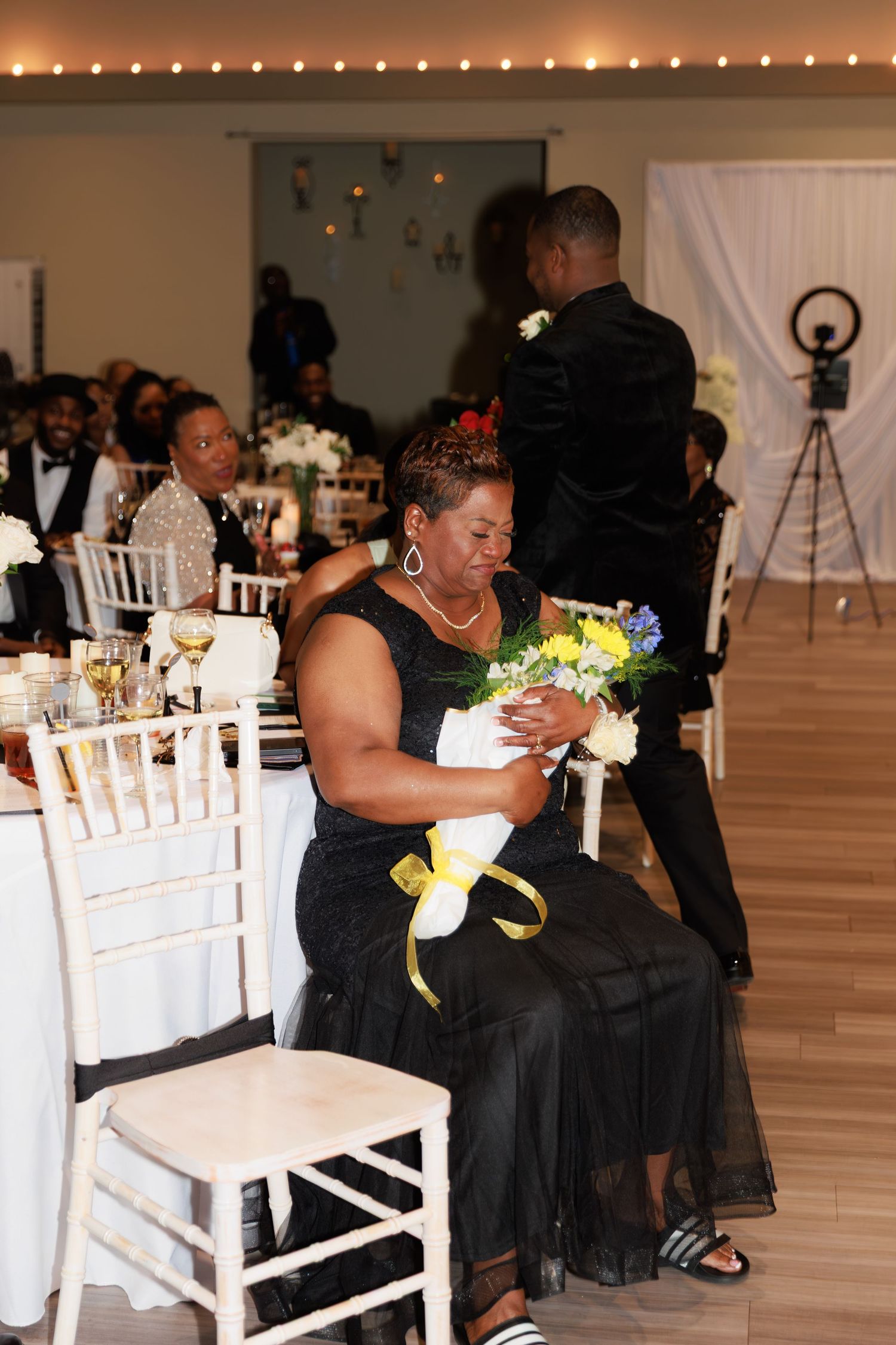 Wedding party members in black dresses and formal attire celebrate at decorated reception tables.