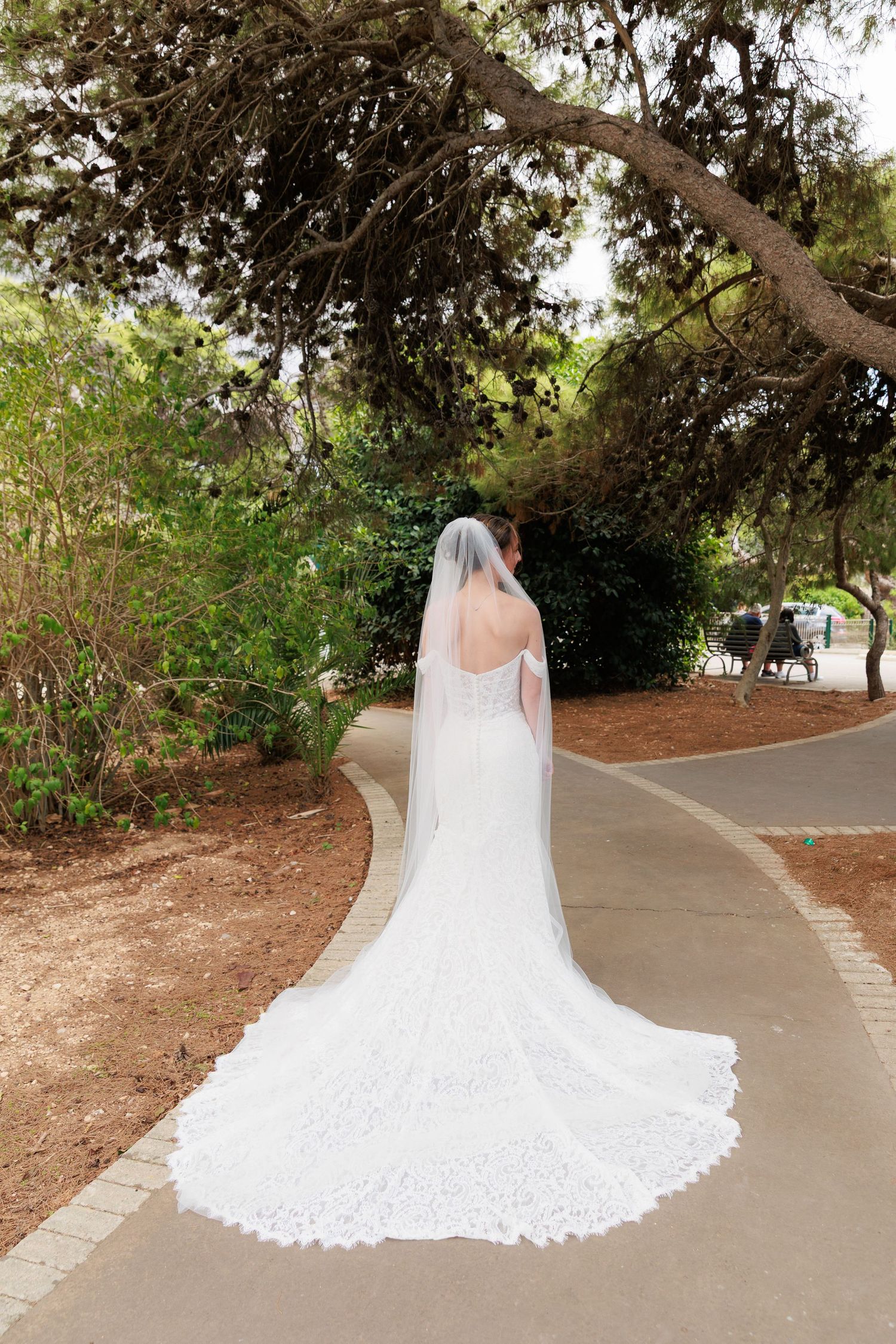 A bride in a flowing white wedding dress with a cathedral-length train stands on a curved garden pathway.