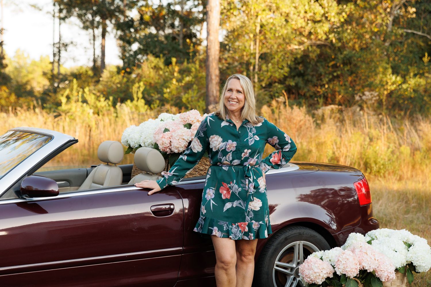 A burgundy convertible car parked in a golden field at sunset with white floral arrangements.
