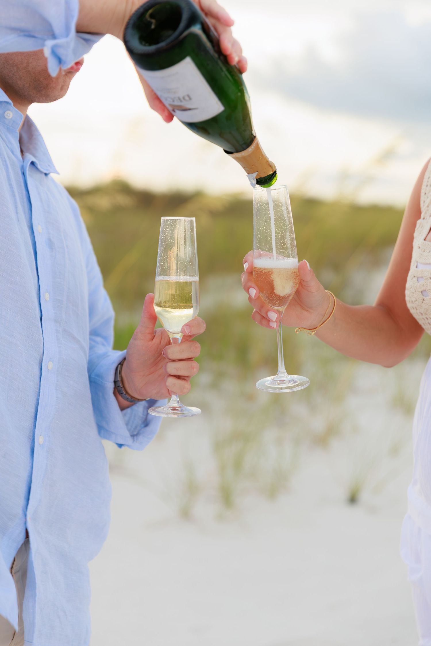 Hands pouring champagne into glasses on a beach with sand dunes in the background.