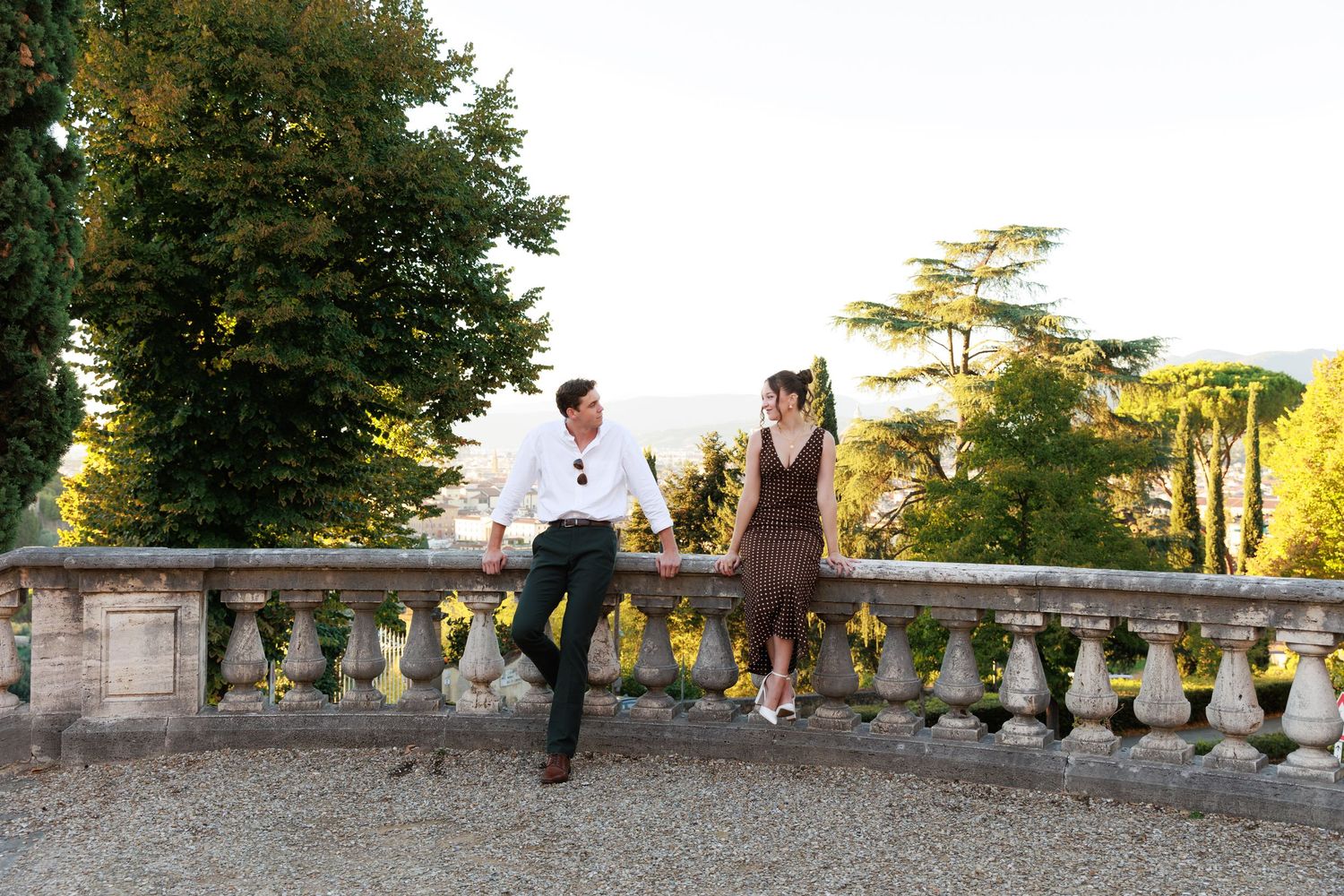Couple sitting on stone balustrade at elegant Italian villa garden with lush trees and scenic views.