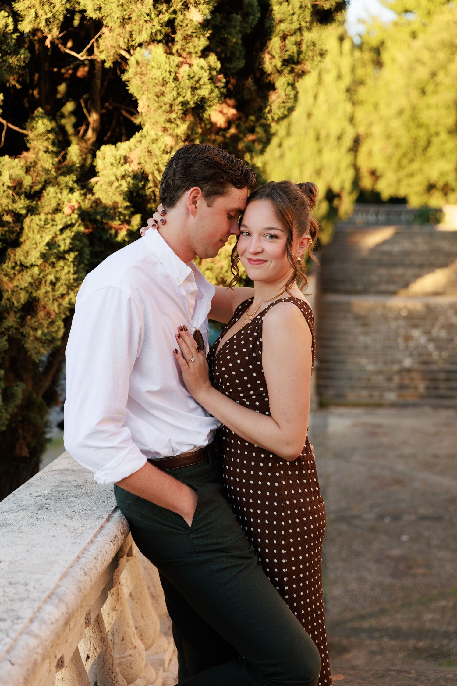Couple embracing tenderly at golden hour near stone balustrade surrounded by lush green trees.