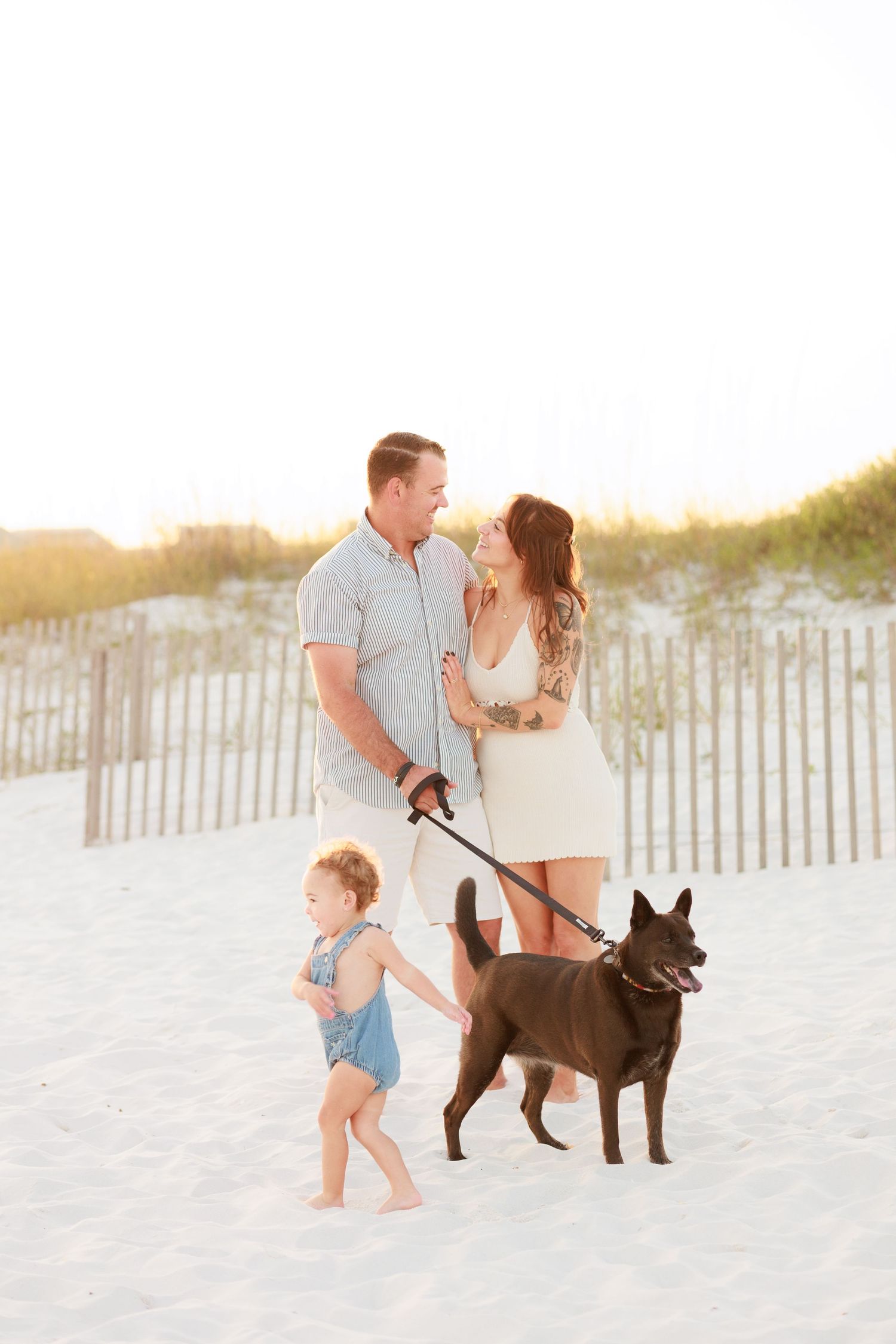 A family with a toddler and dark dog enjoy a sunny beach walk near sand dunes.