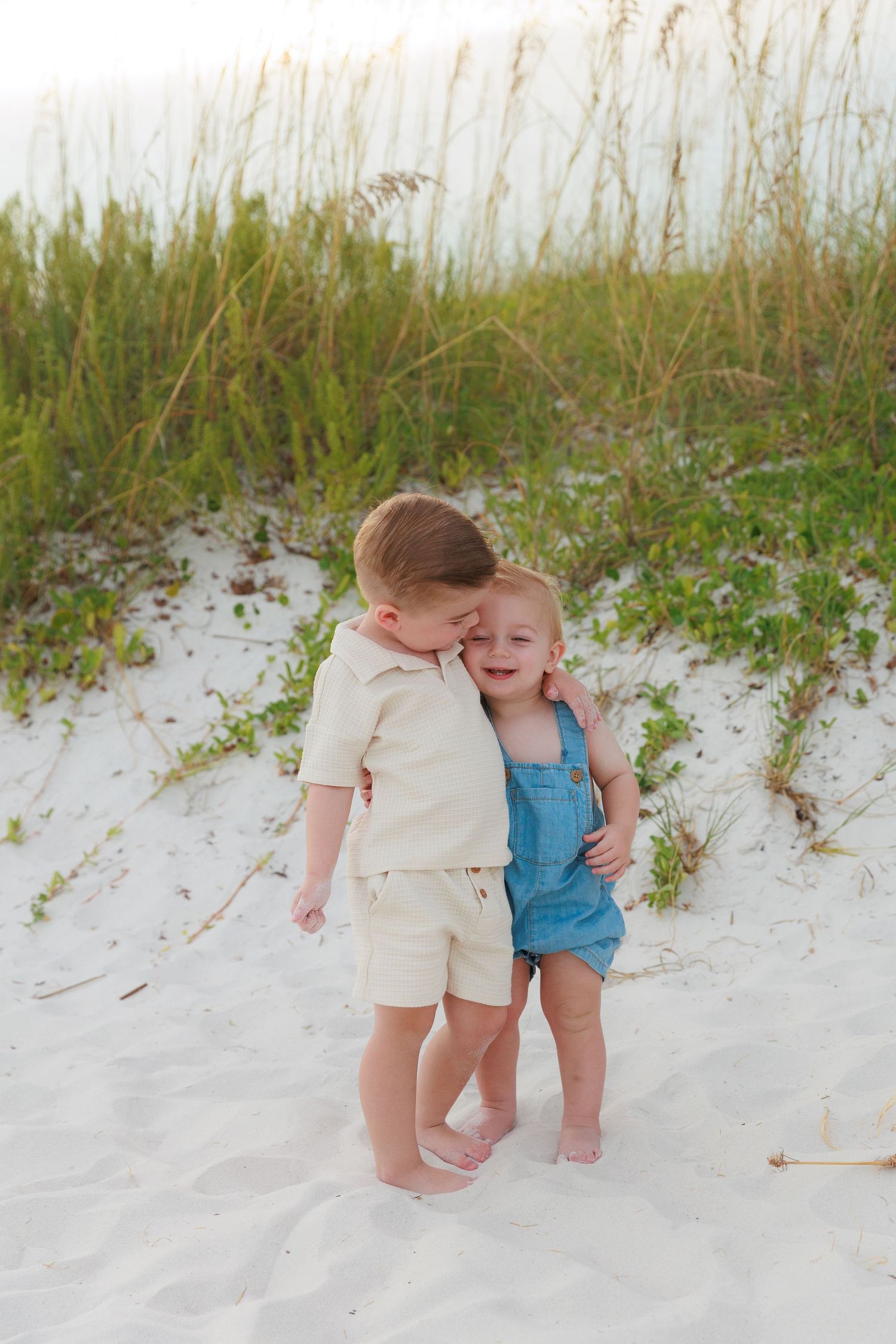 Two young siblings hug on white sand beach dunes, boy in cream outfit kisses girl in blue overalls.