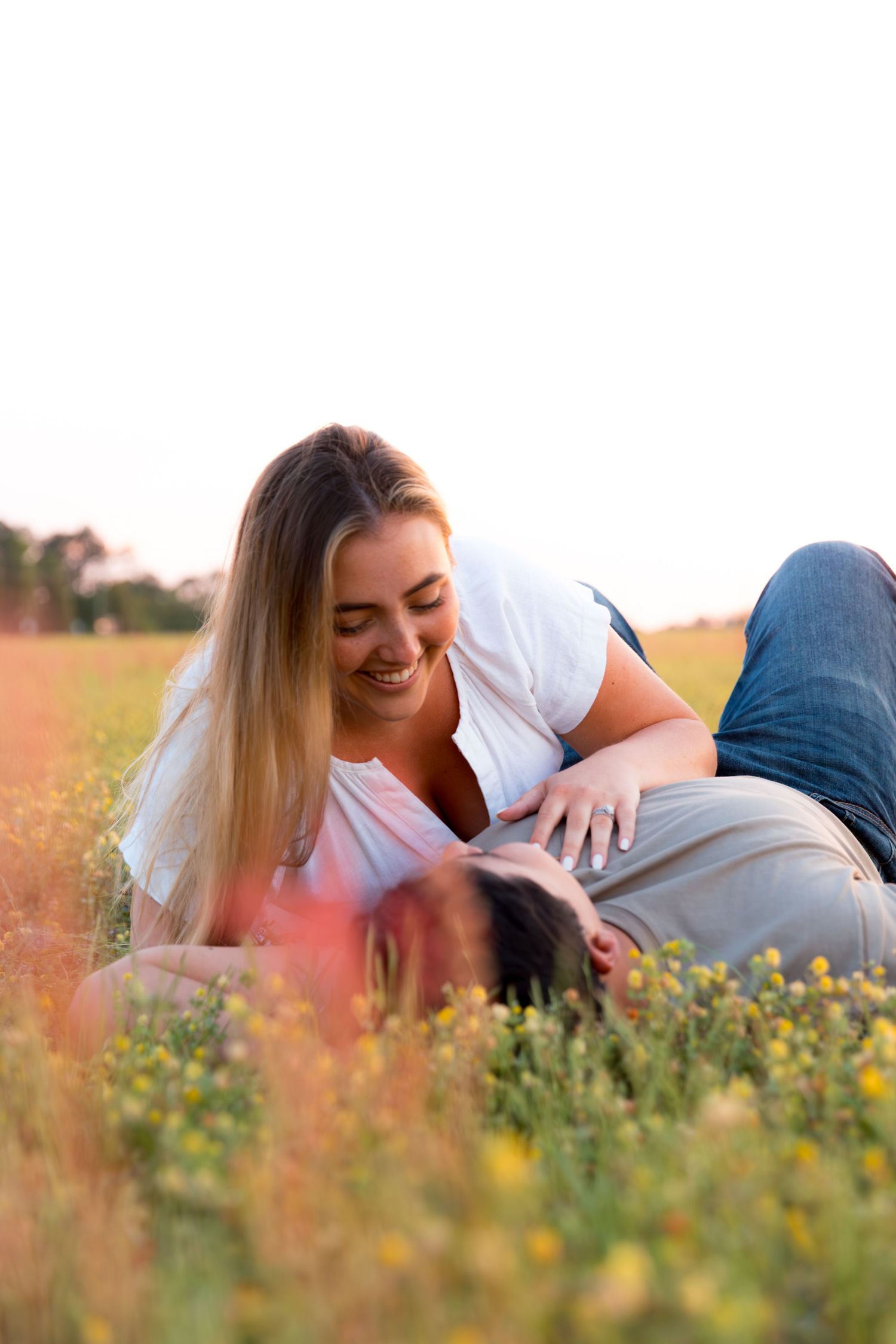 Smiling woman with long blonde hair lying on a man in a sunny wildflower field during golden hour.