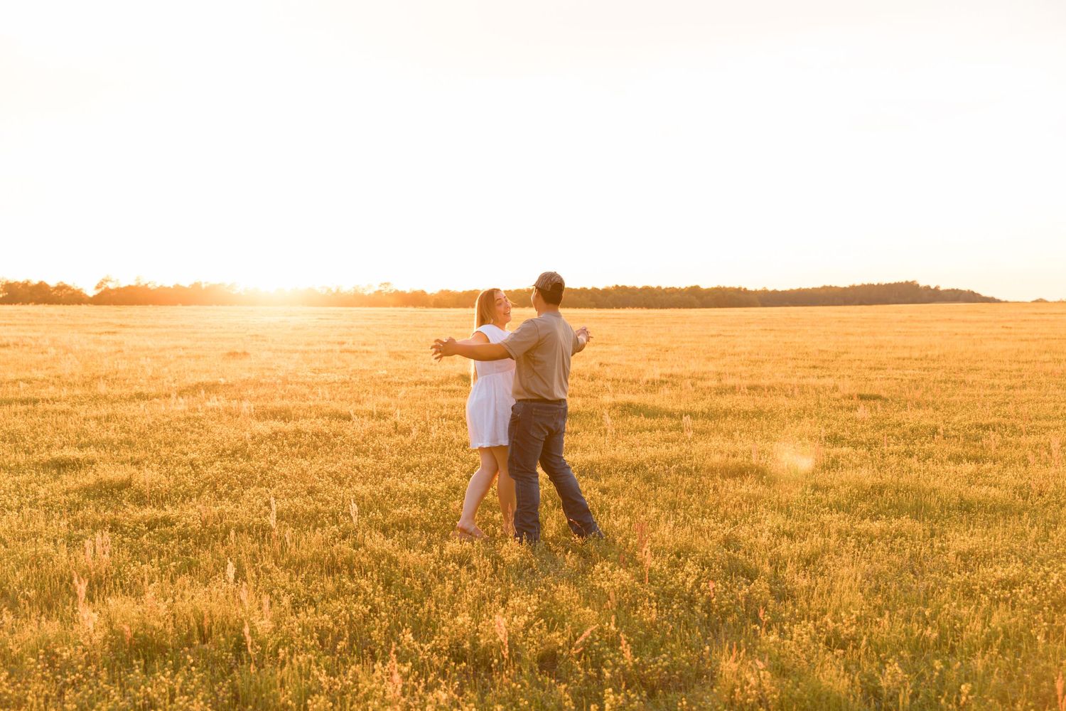 Couple embracing in a golden sunlit field at sunset, facing the horizon with warm light glowing across the meadow.