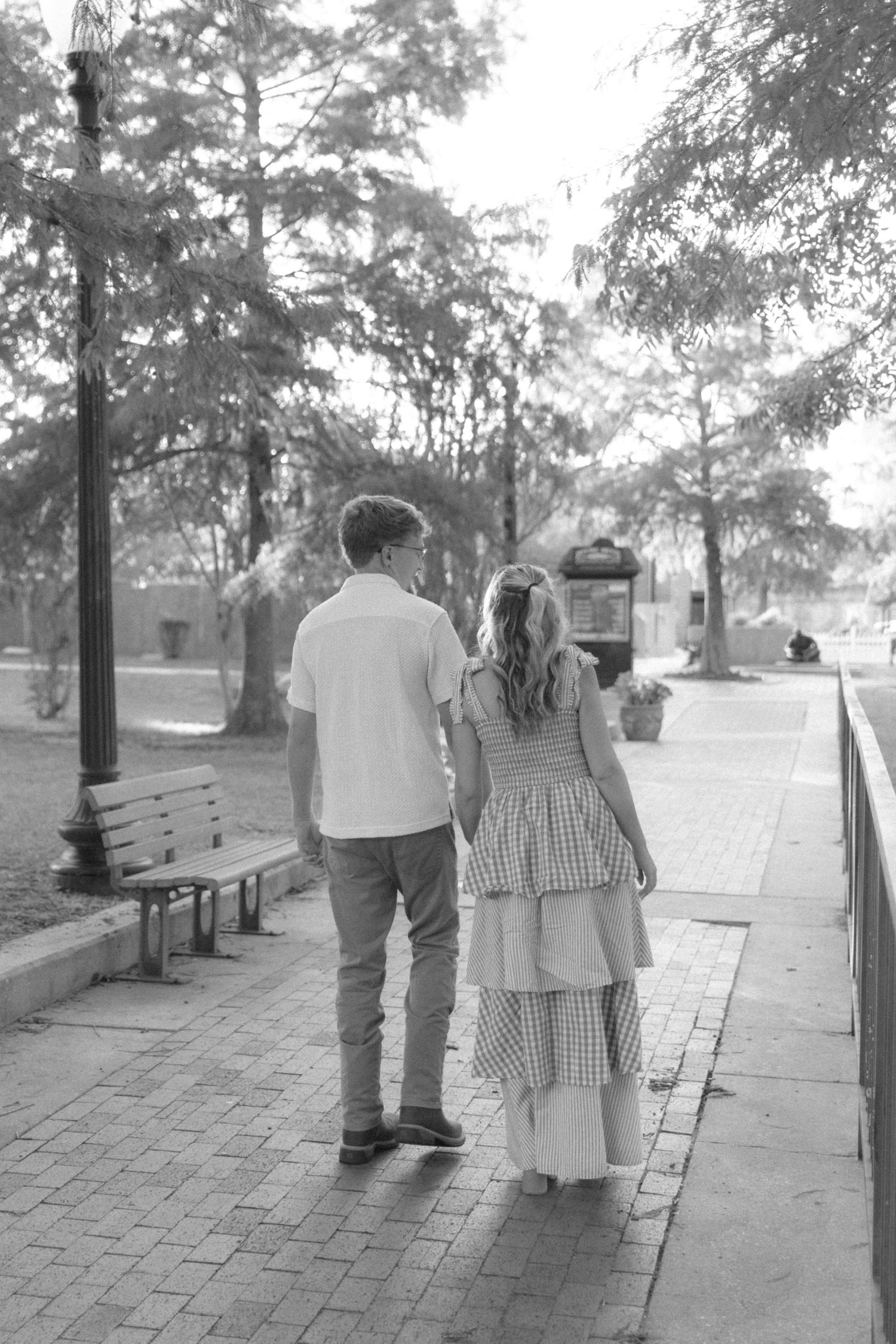 Black and white photo of a couple walking hand in hand along a park sidewalk lined with trees and benches.