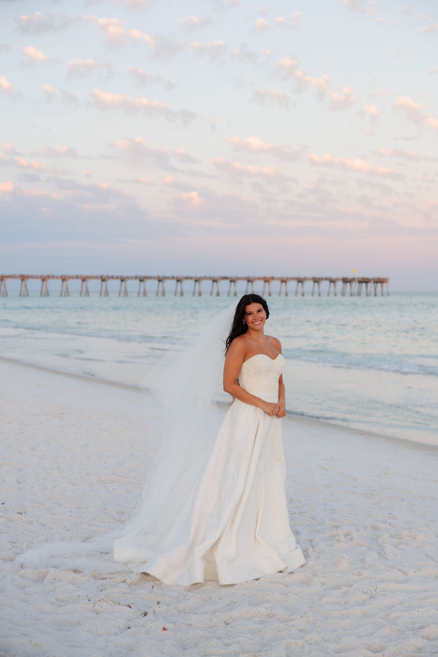 Smiling bride in strapless white gown stands on beach at sunset near pier.