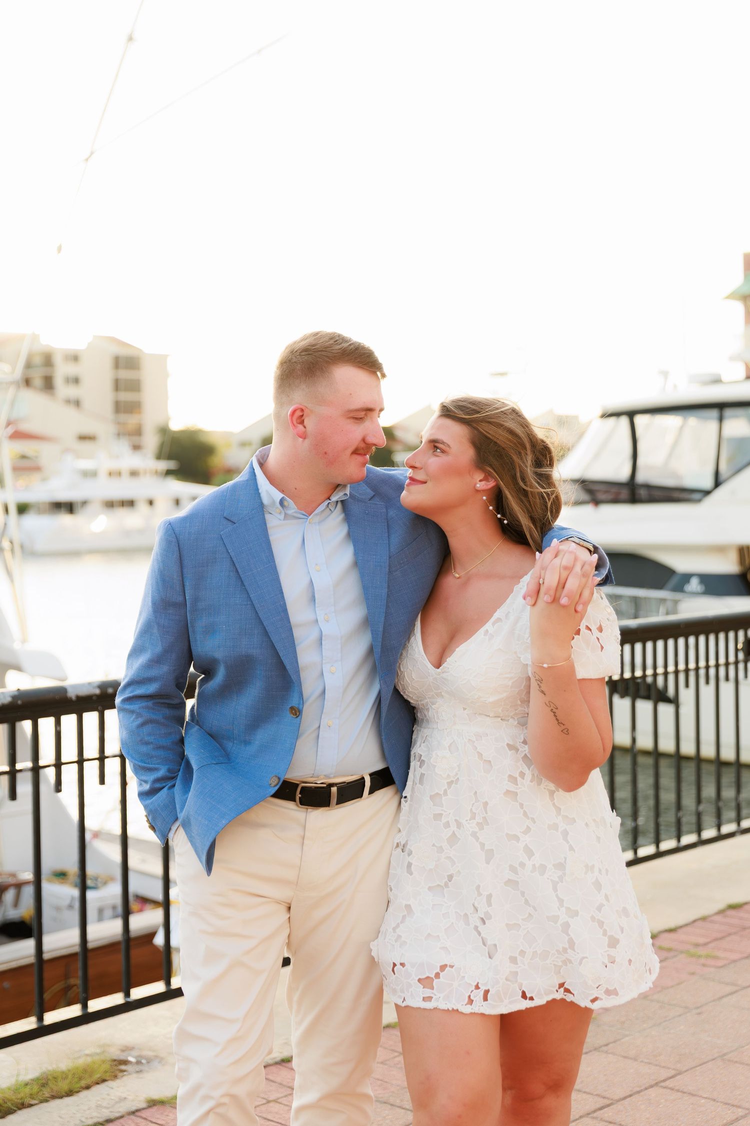 Couple walking arm in arm along a marina boardwalk, man in blue blazer and woman in white lace dress.