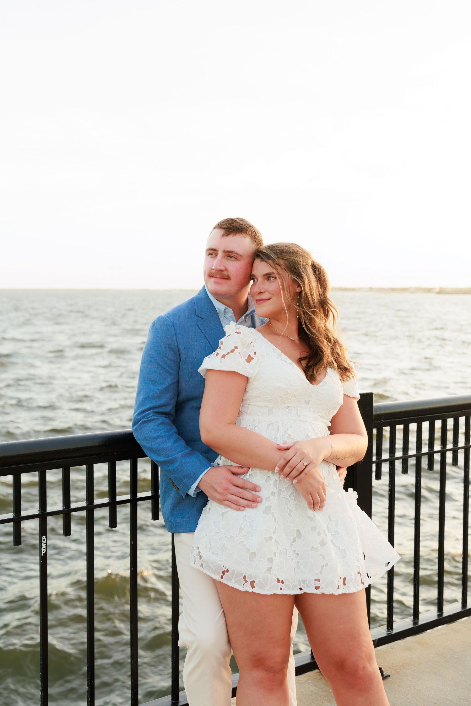 Couple embracing on waterfront pier, man in blue blazer and woman in white lace dress, ocean backdrop.
