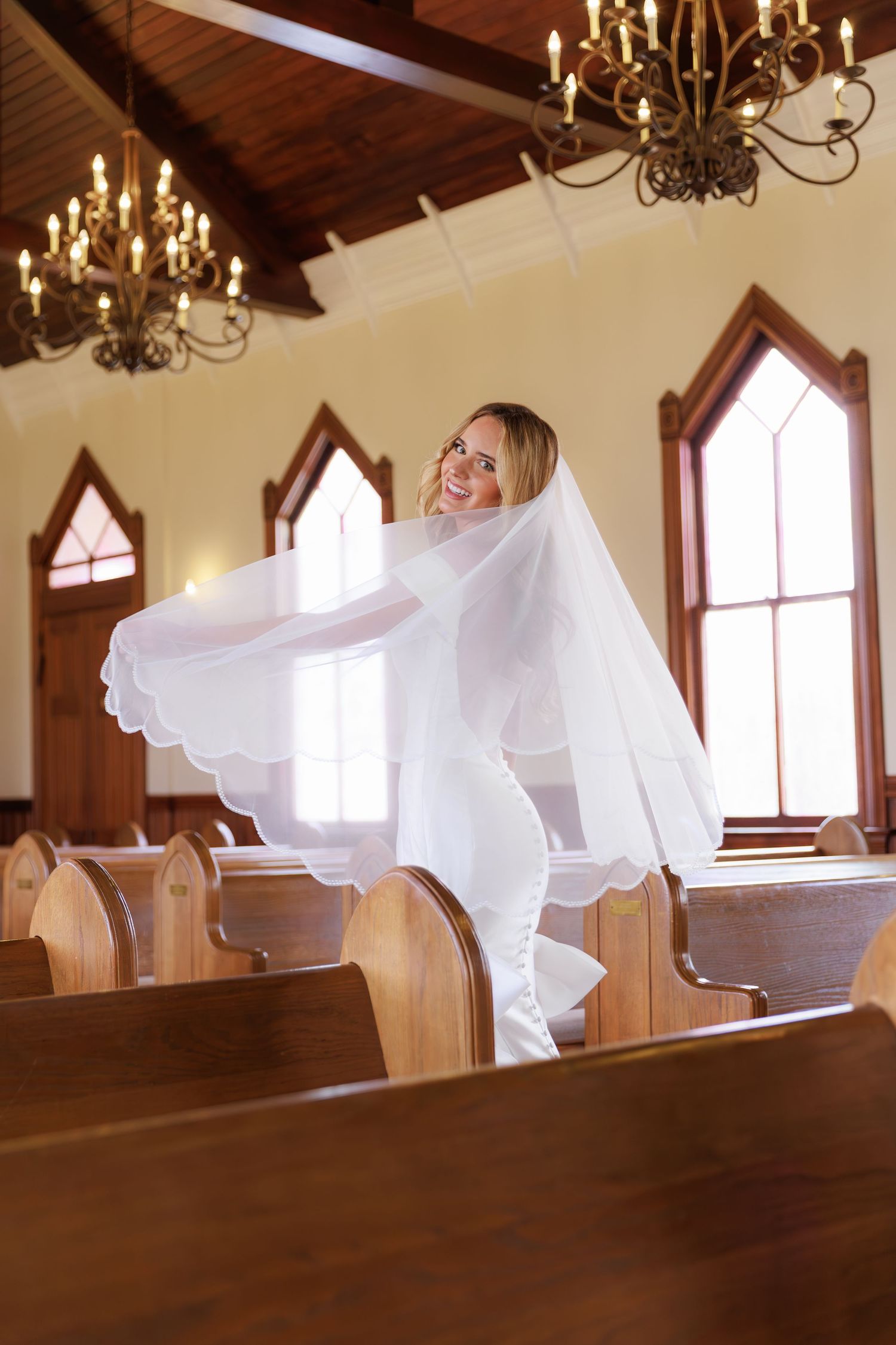 A flowing white wedding dress and veil create an ethereal scene in a church sanctuary with wooden pews and chandeliers.