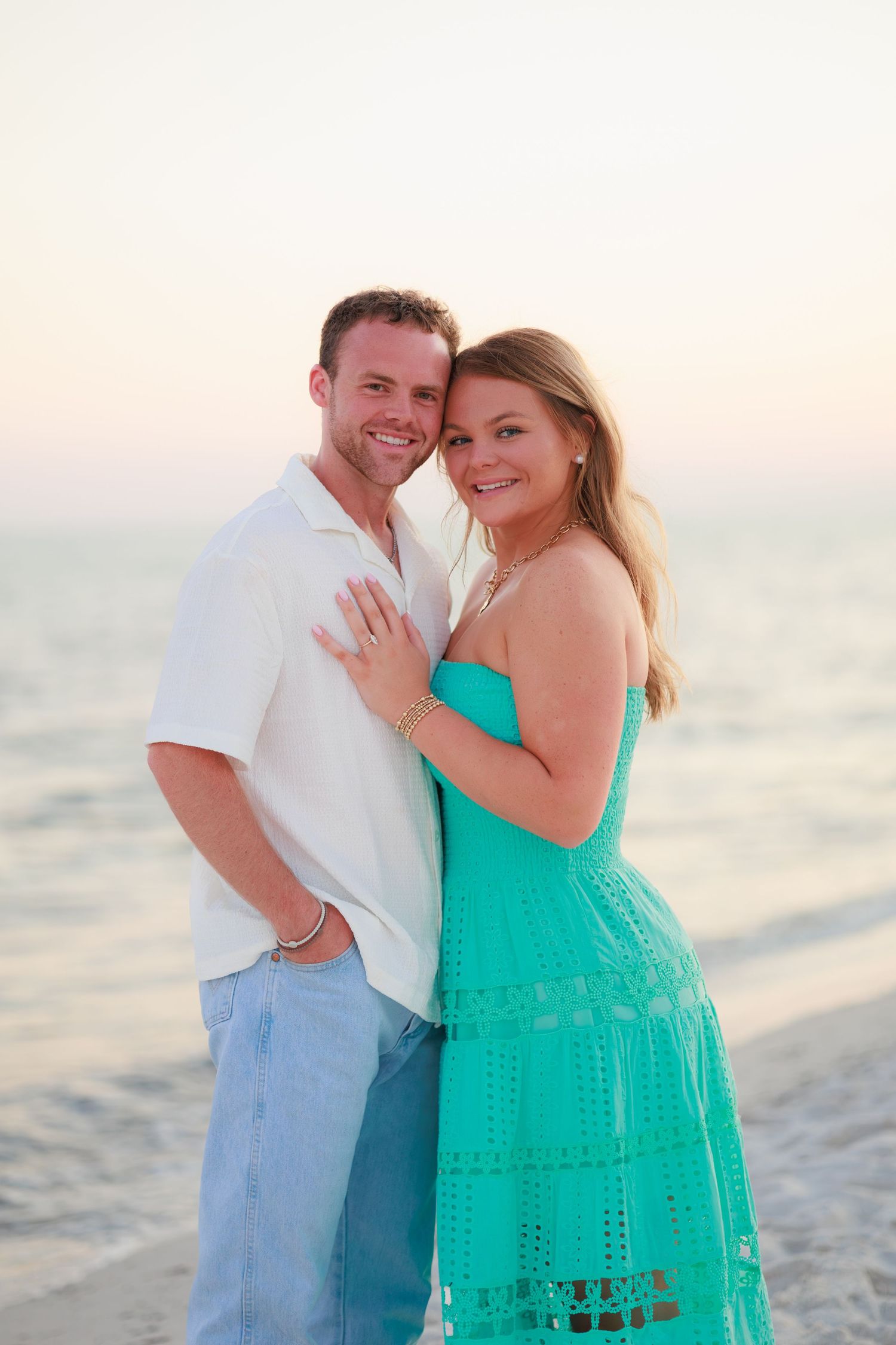 A couple shares a romantic moment on the beach at sunset, with the woman wearing a bright turquoise sundress.