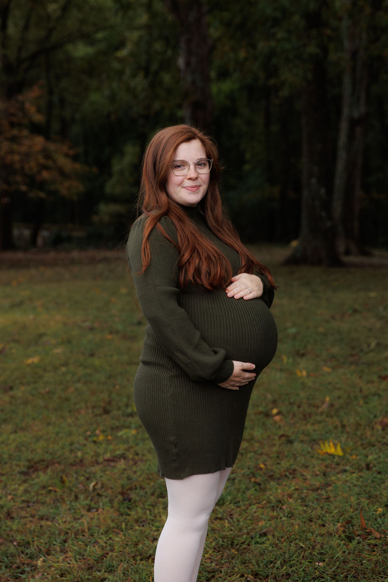 A pregnant woman in an olive green sweater dress stands smiling in a wooded outdoor setting during fall.