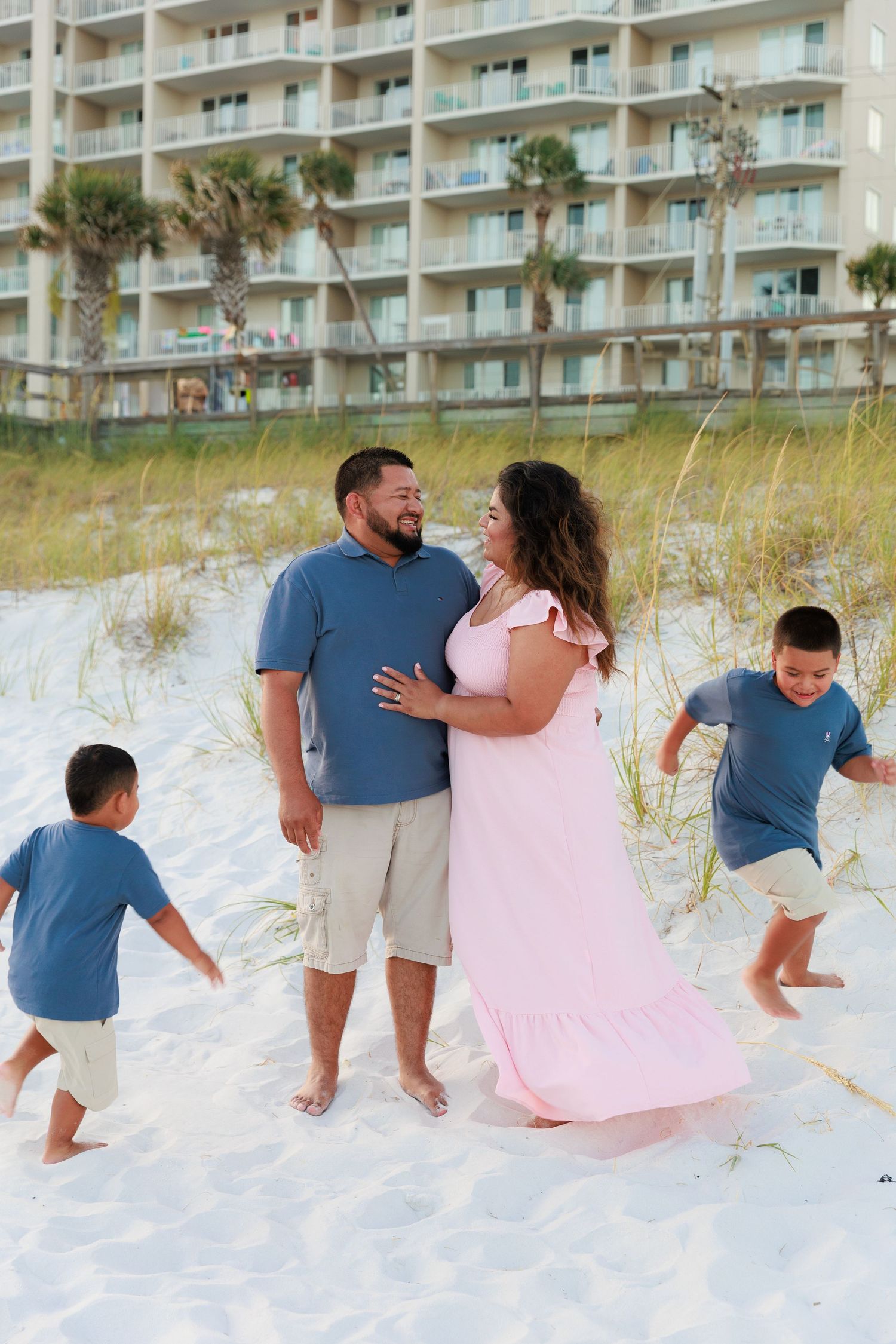 A family shares tender moments on a white sandy beach with beachfront condos in the background.