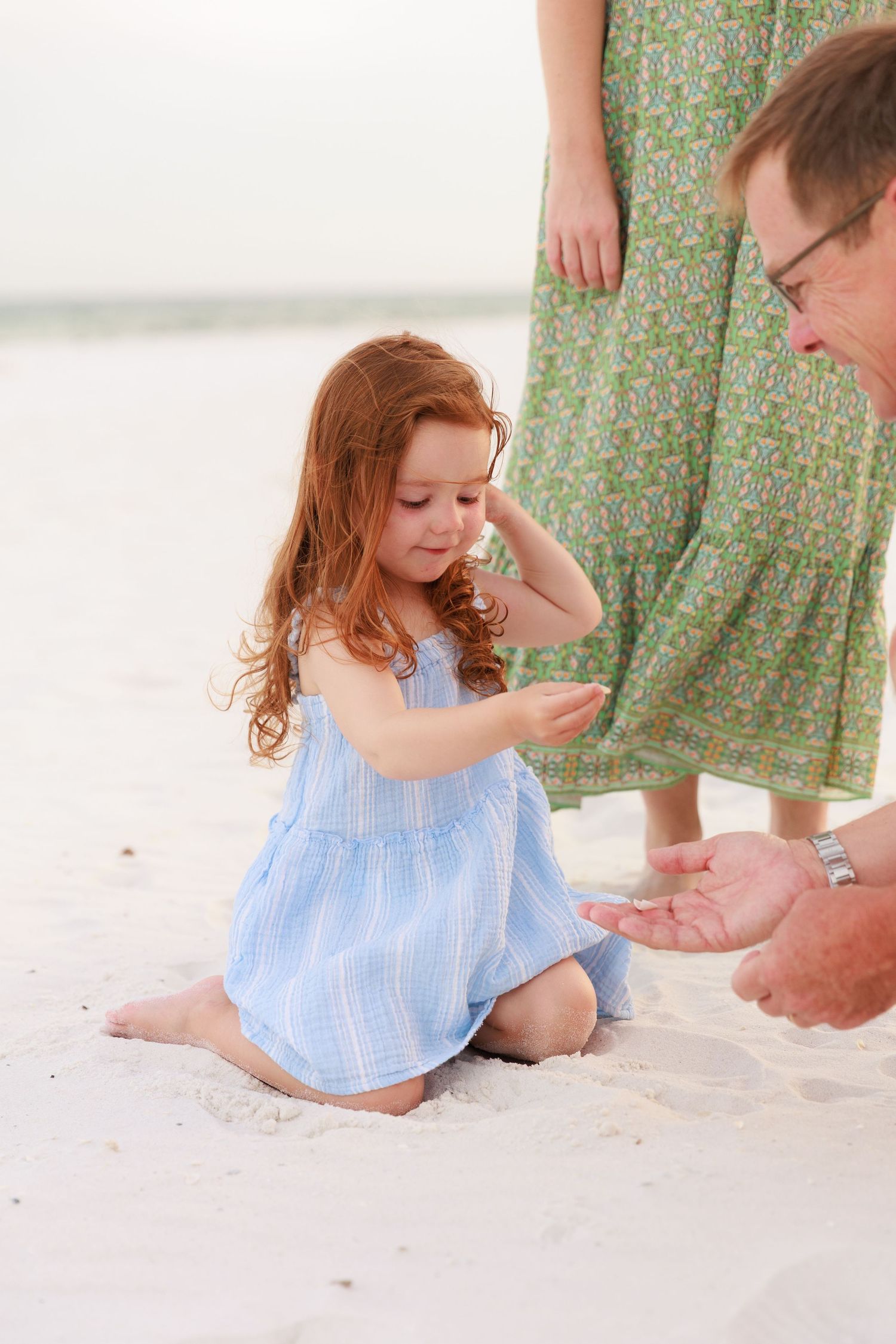 Young child in a light blue dress plays with sand on a white surface while an adult assists.