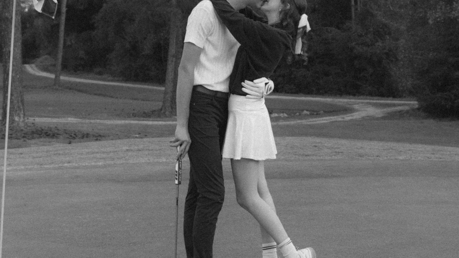 A black and white photo shows two people embracing on a road, with one wearing a white dress and the other in dark clothing.