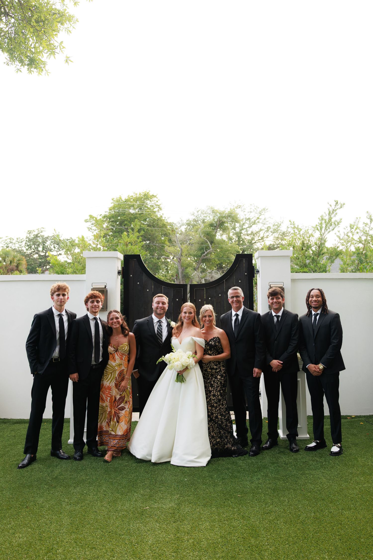 Wedding party poses outdoors on green lawn in front of black iron gate, bride in white gown holding bouquet.