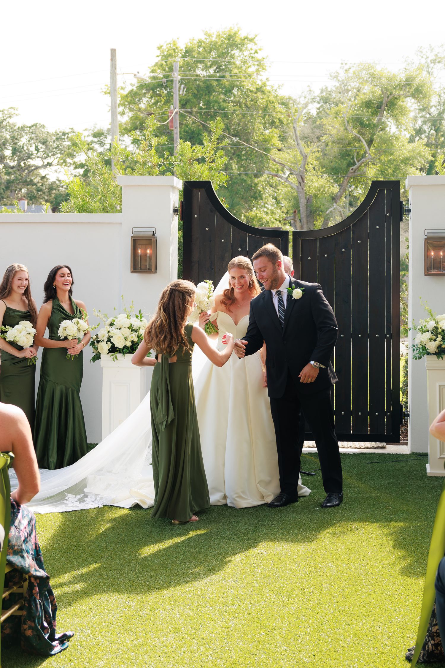 Bridesmaid assists bride during outdoor wedding ceremony with groom, green-dressed bridesmaids, and floral arch backdrop.