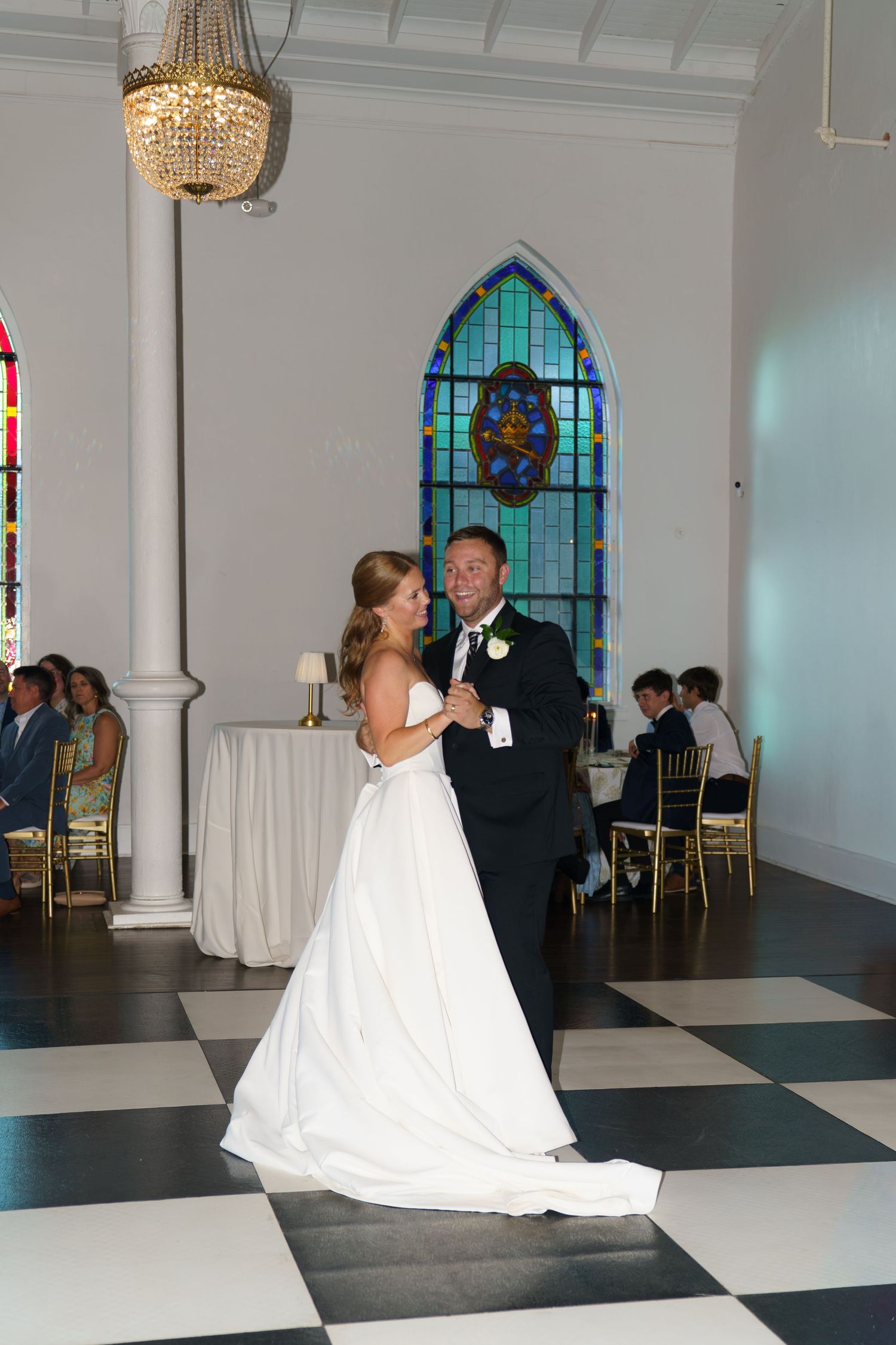 Bride and groom share their first dance on a black and white checkered floor in an elegant chapel with stained glass windows.