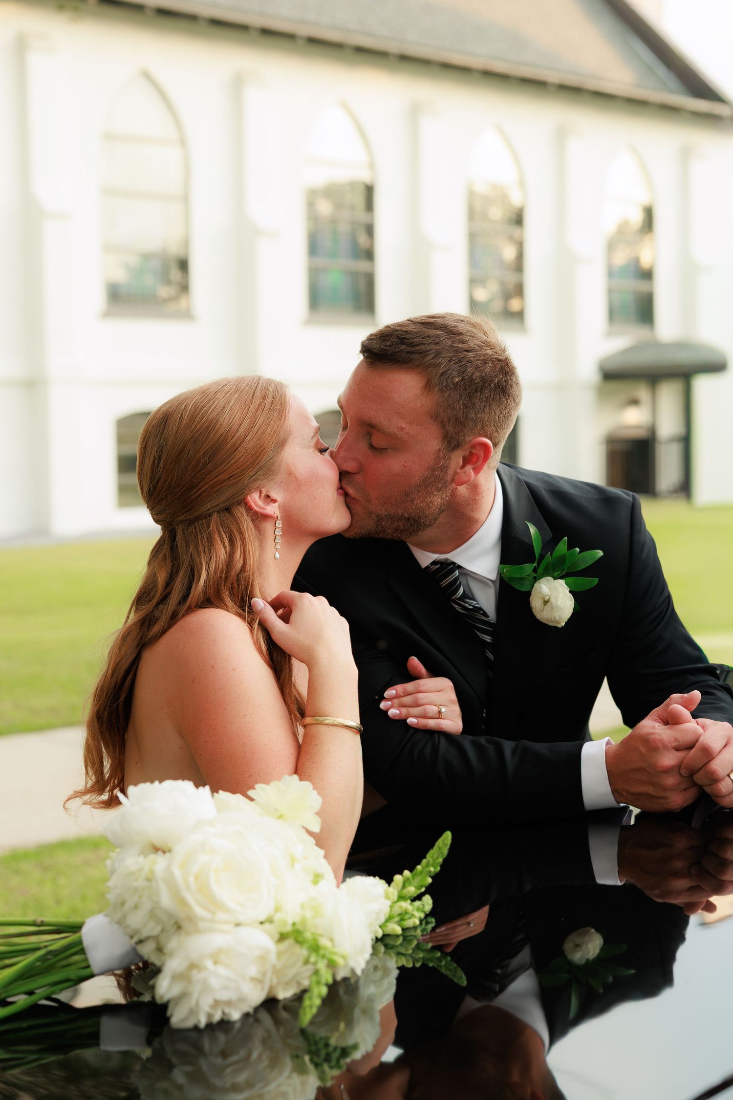 Bride and groom share a romantic kiss, bride holding white floral bouquet outside a white building.