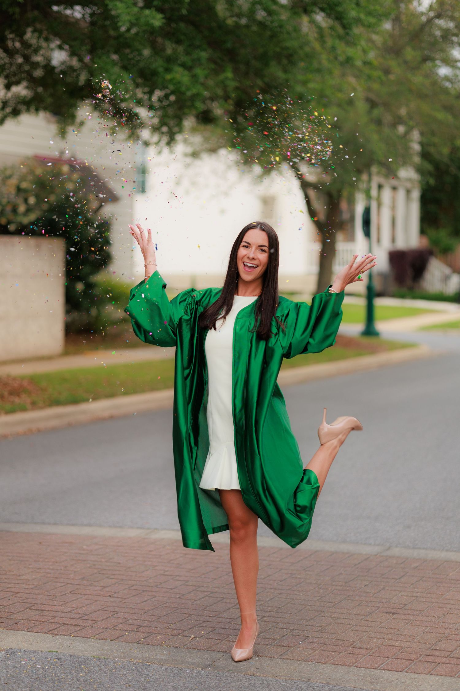 Joyful graduate in green cap and gown tossing confetti while balancing on one leg outdoors.