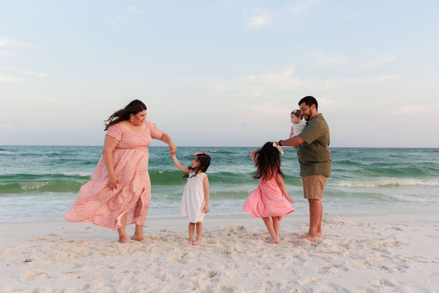 Family of four enjoying a beach day, with parents and two young girls in pink dresses playing at the ocean's edge.