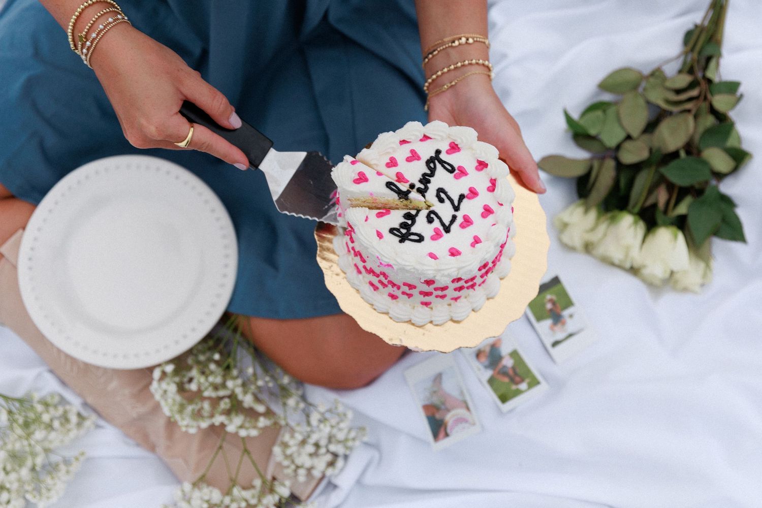 Overhead view of hands holding a decorated pink and white birthday cake on picnic blanket.