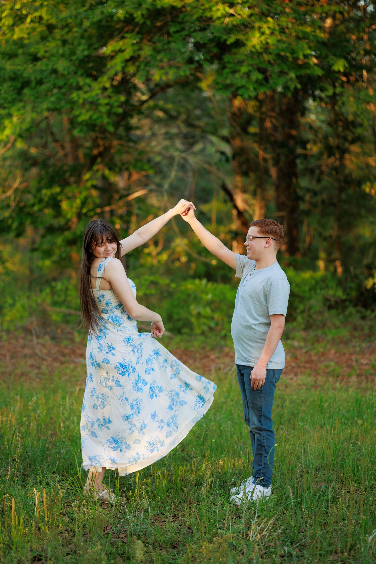 A couple dances together in a green meadow, the woman wearing a white floral dress as the man gently spins her hand.