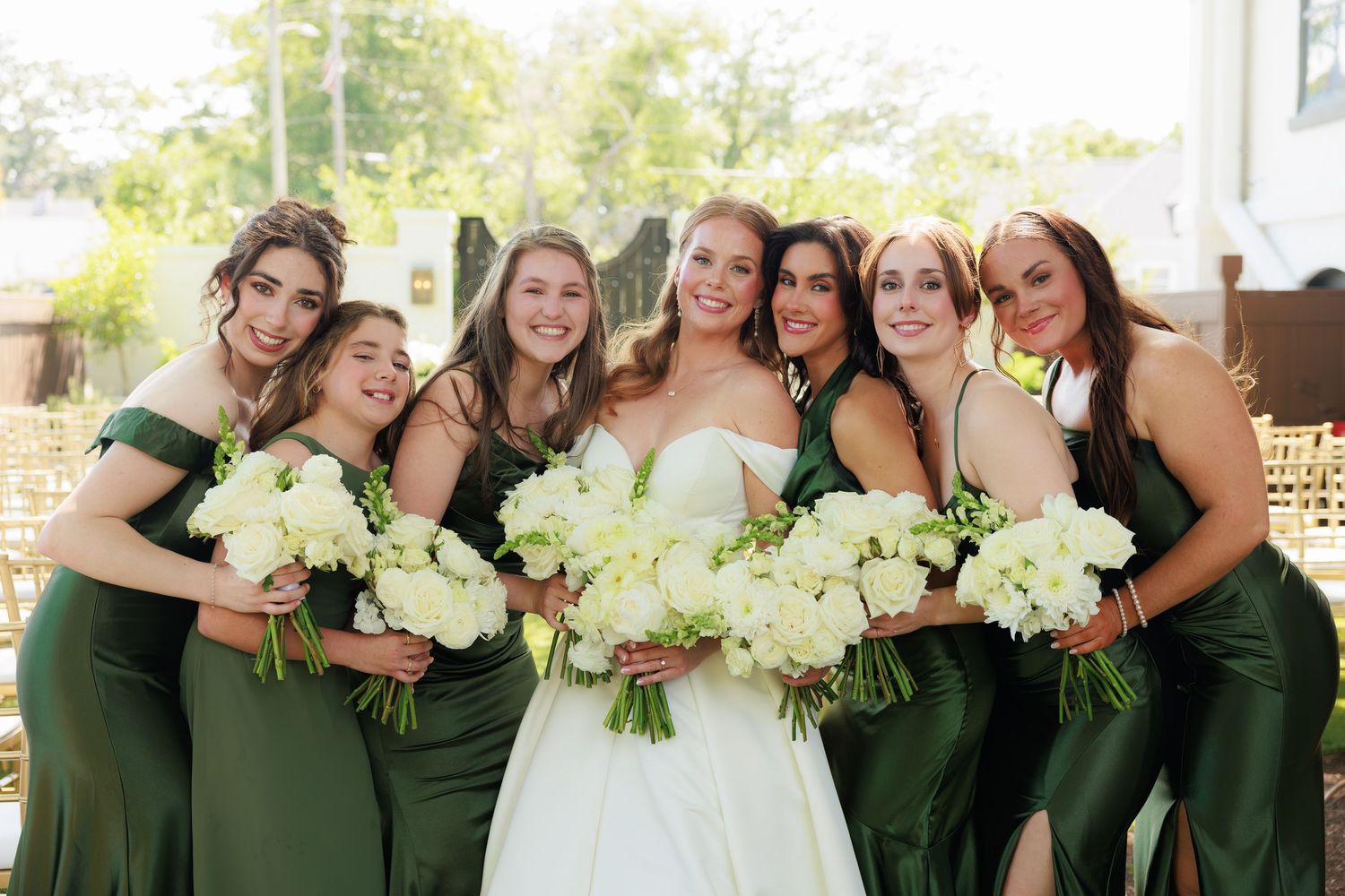 Bride in white gown poses with six bridesmaids in emerald green dresses holding white floral bouquets outdoors.