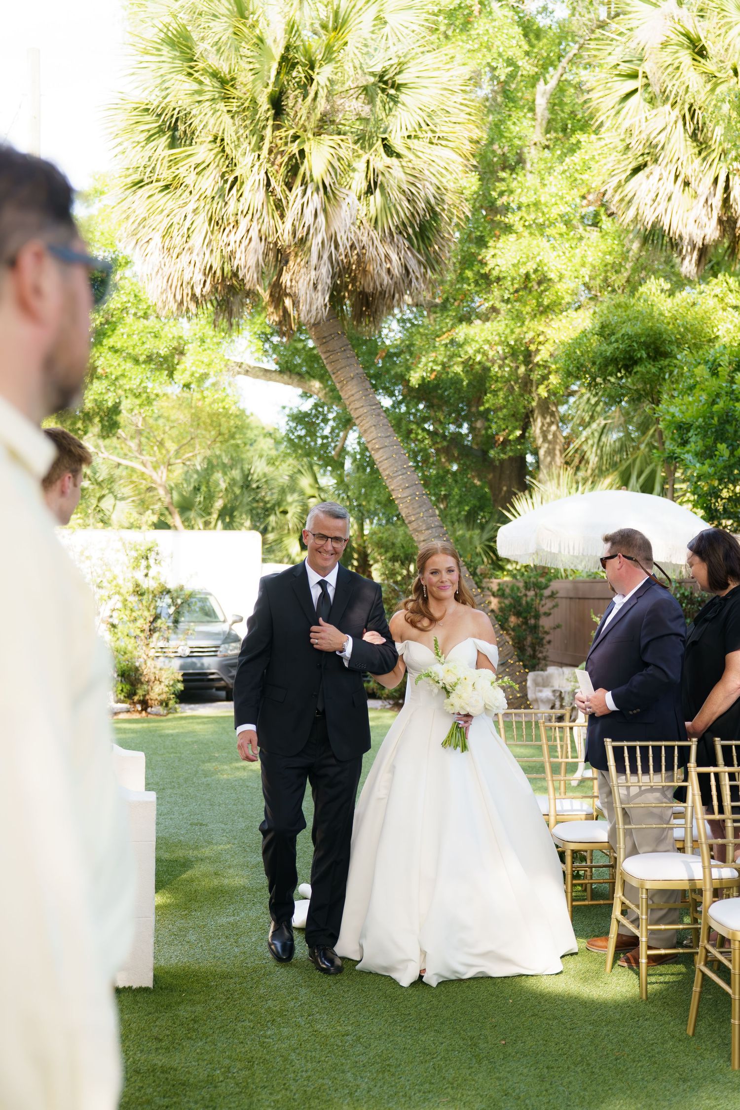 Bride walks down outdoor aisle with escort, wearing off-shoulder gown, holding bouquet, palm trees in background.