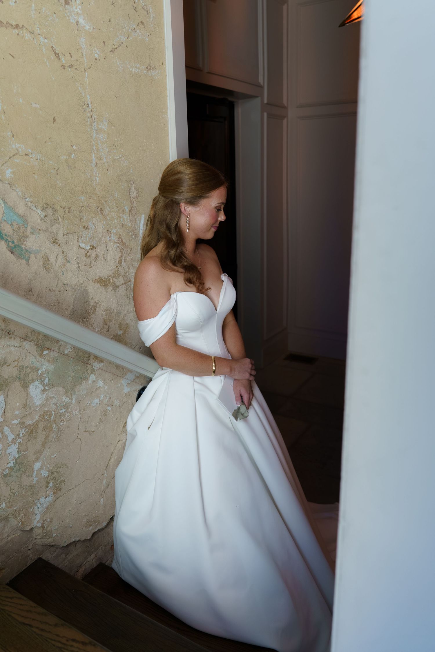 Elegant bride in off-shoulder white ball gown standing on staircase in vintage venue.